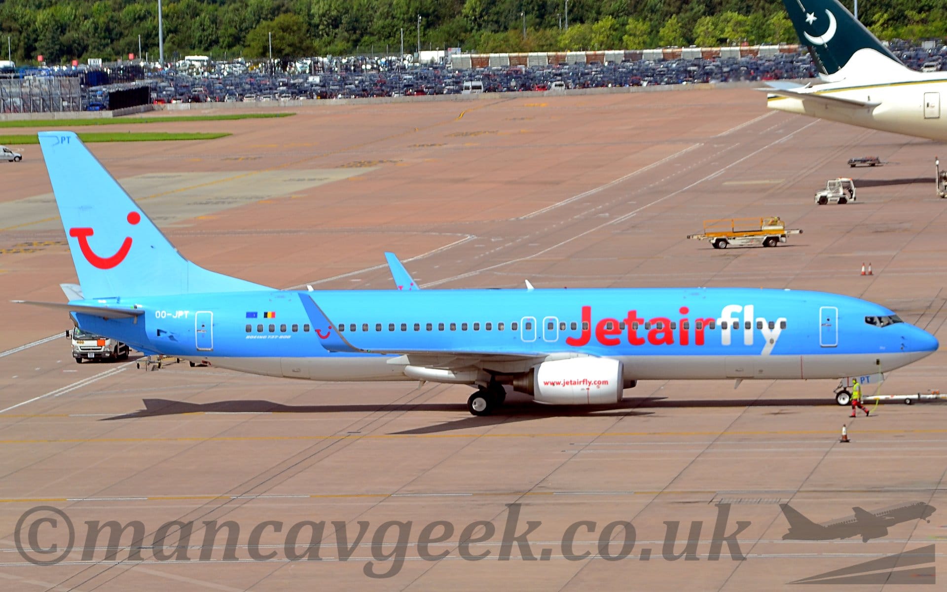 Side view of a twin engined jet airliner facing to the right but being pushed back to the left by an out-of-shot tug, attached to the nosewheel by a long white pole. The plane is mostly blue, with a white belly. There are red "JetAir" titles on the forward fuselage, followed by "white" fly" titles, and the white registration "OO-JPT" on the upper rear fuselage. The blue tail has a red logo that looks to be a cross between a winking smiley face, and the letters "T", "U", and "I" - this logo also appears on the upturned wingtips. The white engine pods slung under the wings have the red web address "www.jetairfly.com". Grey concrete apron fills the foreground and background, with the white rear fuselage and green and white tail on the right of the frame. A large, ground-level car park is right at the back of the frame, with trees right beyond that.