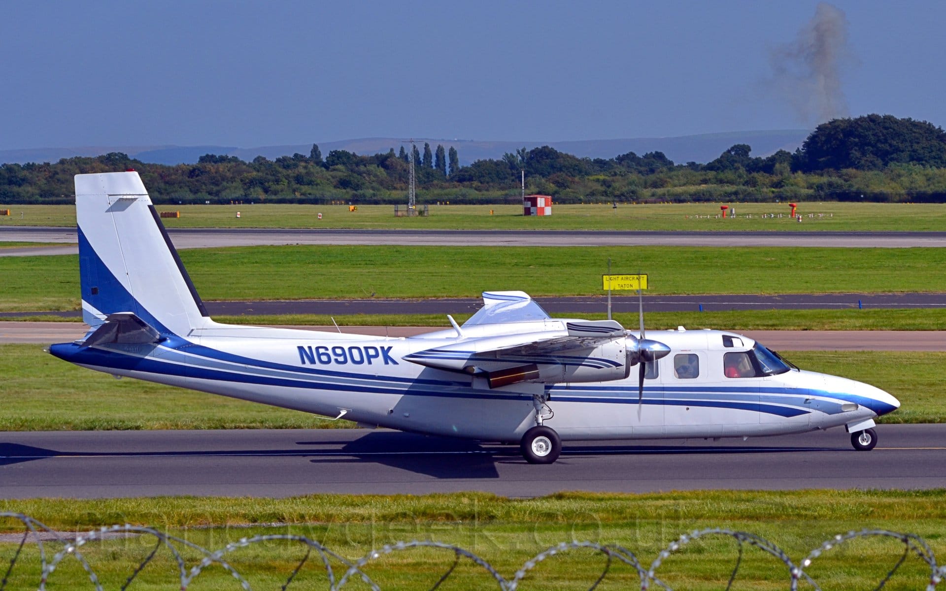 Side view of a high-winged, twin engined BizProp taxiing from left to right. The plane is largely white, with a 2-tone blue stripe running along the body from under the nose, and sweeping up into the lower parts of the tail. The registration N690PK is on the rear fuselage. Green grass fills the bottom of the frame, lining each side of the taxiway, with more grass in the background, leading up to trees in the distance under blue sky.