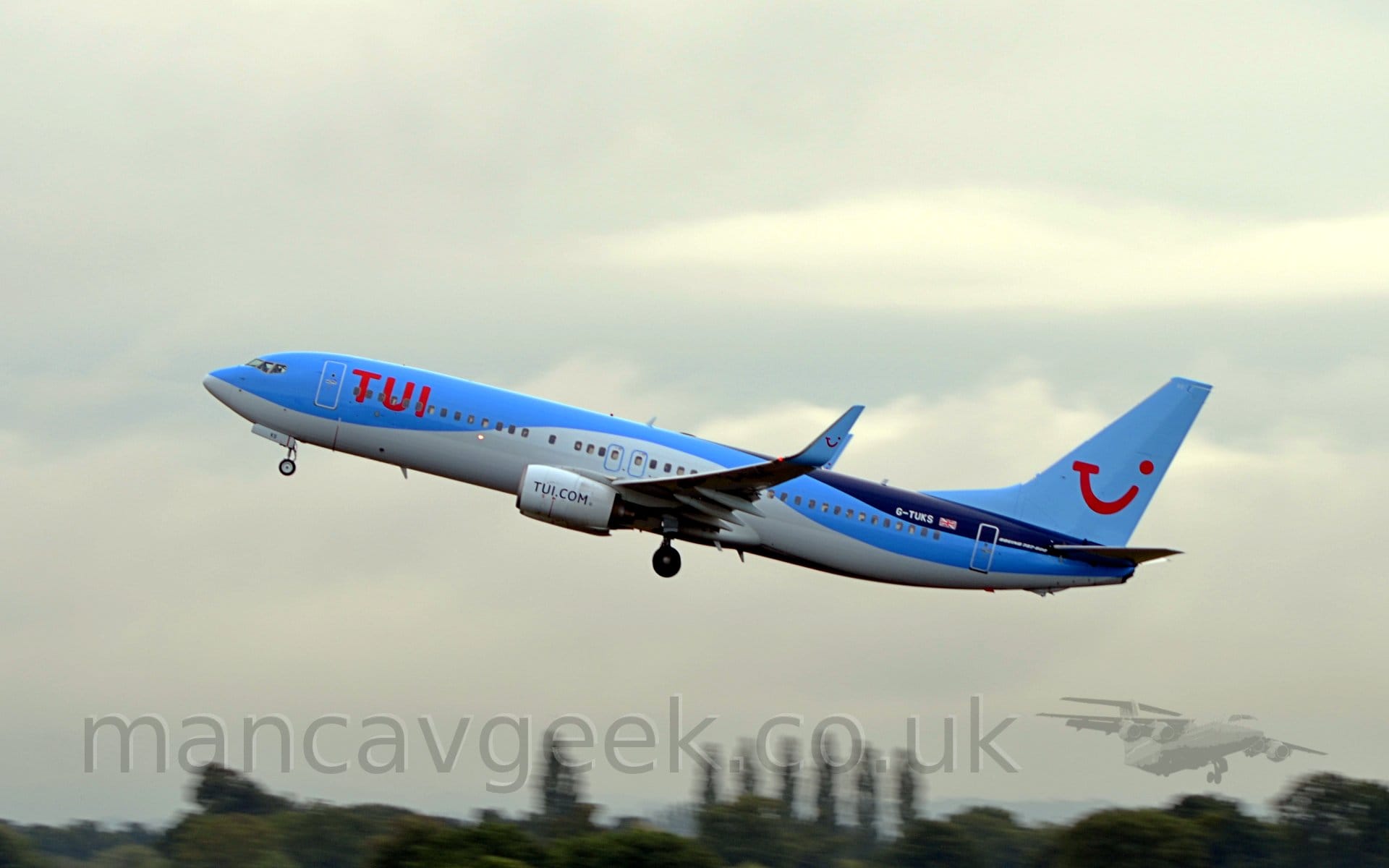 Side view of a twin engined jet airliner flying from right to left at a very low altitude, with undercarriage lowered, flaps extended from the rear of the wings, and the nose raised sharply, suggesting it has just taken off. The plane is light blue on top and white underneath, the white section swooping up over the wings and back down again, with a dark blue section on top of the rear fuselage, under the tail. There are large red "TUI" titles on the forward fuselage. The white registration "G-TUKS" is on the dark blue section of the rear fuselage, next to a small British flag. The tail is light blue, with a large red logo that looks like a red winky face, but is made up of the letters "T", "U", and "I", a smaller version of this logo appearing on the blue upturned wingtips. The web address "tui.com" appears on the side of the white engine pods, slung under the wings. Green trees stretch across the bottom of the frame, while blue-grey cloudy skies with fluffs of white fill the rest.