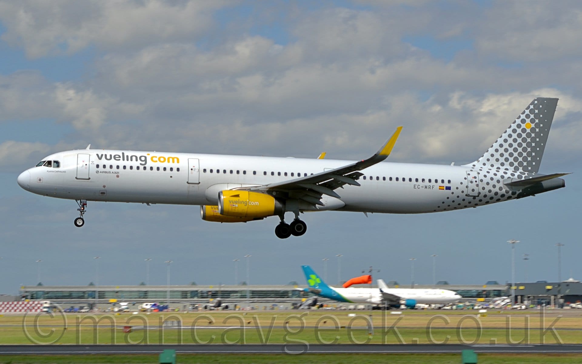 Side view of a twin engined jet airliner flying from right to left at a very low altitude, with undercarriage lowered and flaps extended from the rear of the wings, suggested it is about to land. The plane is mostly white, with grey "Vueling" titles on the upper forward fuselage, with additional yellow "Com" text immediately after. The grey registration "EC-MRF" is on the lower rear fuselage, next to small Spanish and EU flags. The tail-cone and tail start as white with a series of small grey dots, slowly transitioning to grey with white dots, then all grey. A single yellow dot is in the middle of all of these grey and white dots. The engine pods mounted under the wings are a bright yellow, with grey and white "Vueling Com" text. The upturned wingtips are also bright yellow. Bright green grass lines both sides of the runway in the foreground at the bottom of the frame, leading up to a heavily glazed grey terminal building, with a white and blue-green plane in front of it. Blue sky with fluffs of white and grey cloud fill the rest of the frame.