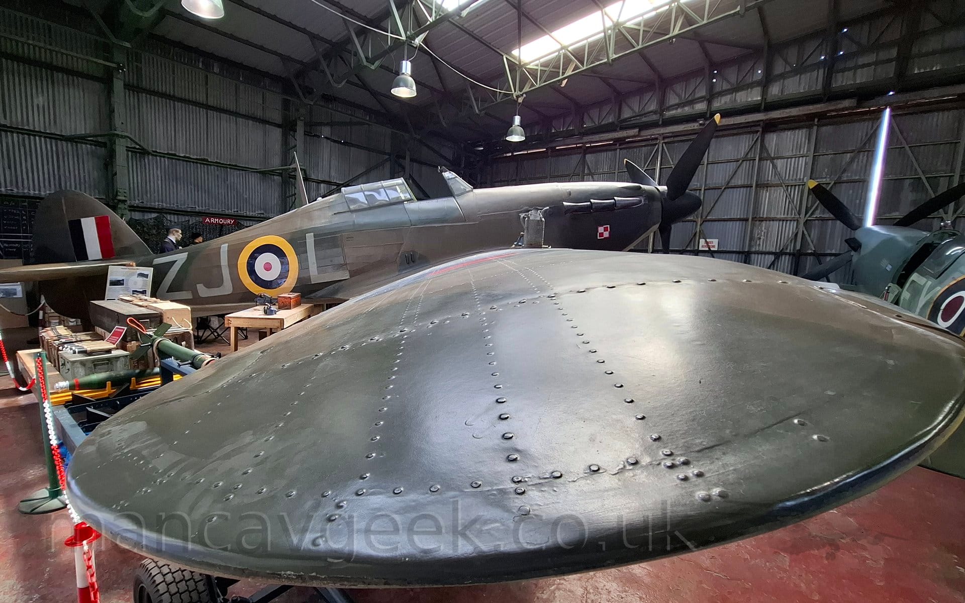 Side view of a single engined, World War 2-era military fighter aircraft in a museum, facing to the left, focusing on a close-up of the right wing. The plane is covered with a brown and olive green camouflage scheme, with a yellow, blue, white, and red roundel on the rear fuselage, with white letters "ZJ" on one side to the rear, and "L" towards the front. There is a blue, white, and red fin flash on the tail, and a Polish military logo (4 squares, 2 white and 2 red, arranged in a square, outlined with contrasting red and white lines) on the engine cowling in the nose. Museum exhibits are scattered around the plane, including a couple of dummies wearing period outlines in the background on the left, and a grey fighter plane on the right. The corrugated metal walls, ceiling, and doors of the old hangar the museum is in fill the rest of the frame, with opaque panels in the ceiling and suspended lights flooding the room with light.
