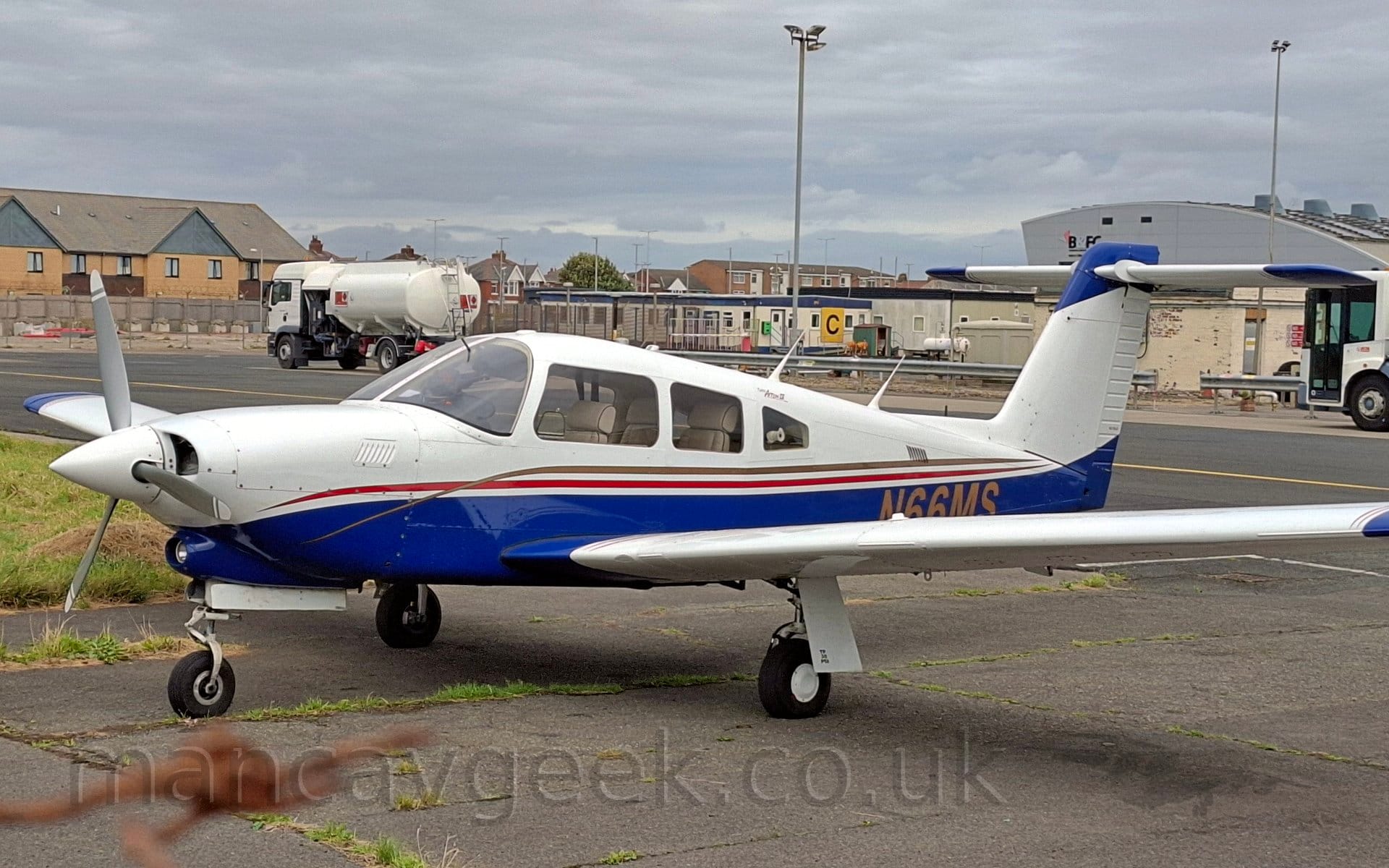 Side view of a single-engined light aircraft with T-tail, parked black tarmac apron facing to the left. The plane is largely white, with a blue lower half, with a red and gold stripe running along the body. The gold registration "N66MS is on the lower rear fuselage, the bottom partially obscured by the wing. A pair of fuel trucks are parked in the background in front of some grey pre-fabricated buildings, with a large, squat, grey building further back on the right, and brown brick housing in the middle and on the left, all under cloudy grey sky with occasional hints of blue.