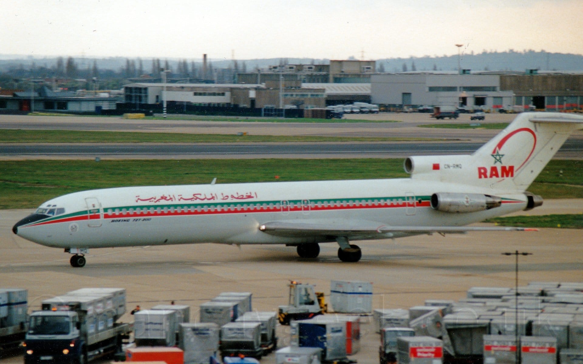 Side view of a 3 engined jet airliner taxiing from right to left. The plane is mostly white, with a green and red cheatline running along the body, covering the passenger cabin windows, and red titles in an Arabic script on the upper forward fuselage. The belly is grey, with black "Boeing 727-200" titles on the forward fuselage. The black registration "CN-RMQ" is on the centre engine inlet above the rear fuselage, next to a Morrocan flag. The other engines are mounted on the sides of the rear fuselage, under the tail. The tail itself is white, with the outline of a 5-pointed green stan with a red swoosh behind it, over red "RAM" text. The foreground is filled with metal luggage containers and vehicles, while the background has large grassed areas either side of a black runway, with a taxiway and several buildings beyond that, with flat white sky over everything.