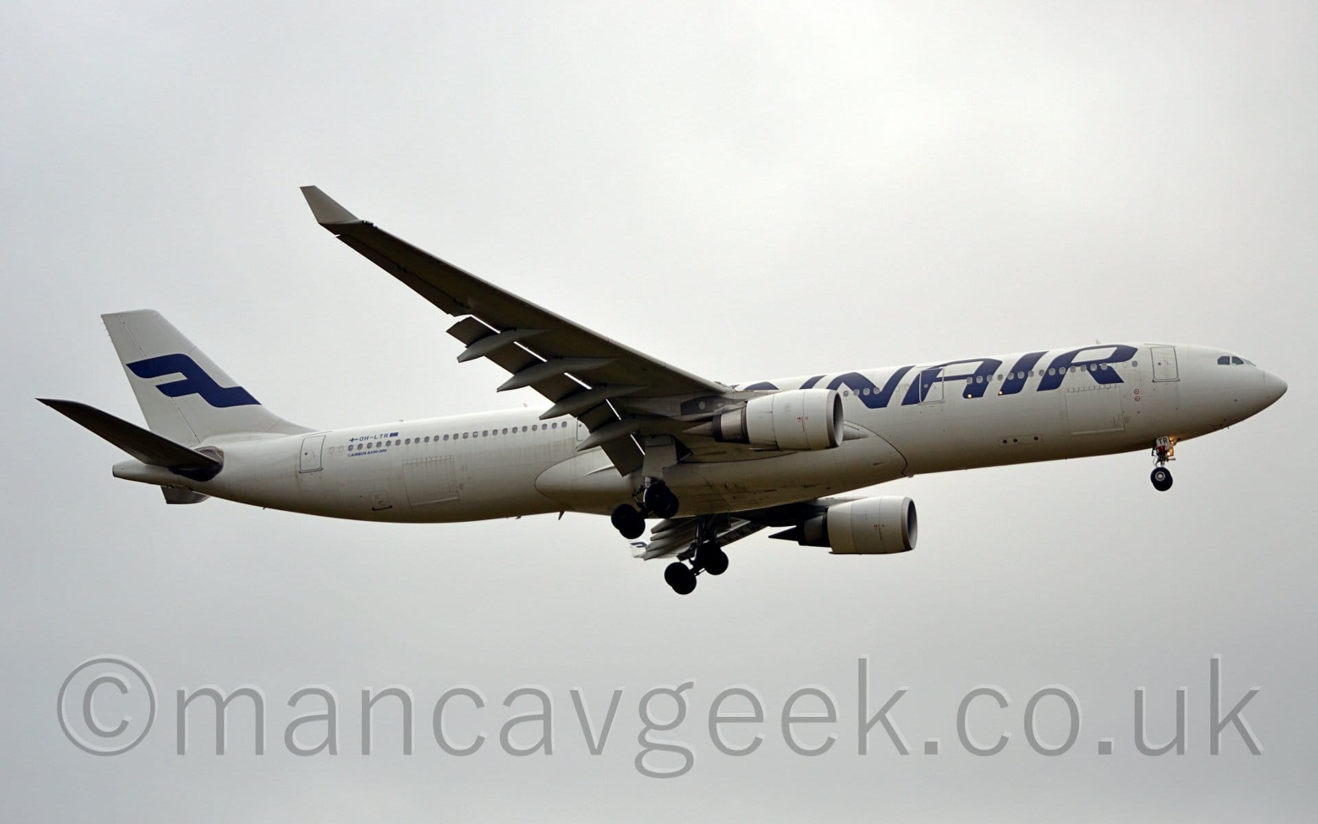 Side view of a twin engined jet airliner flying from left to right at a low altitude with undercarriage lowered, flaps extended from the rear of the wings, suggesting it is about to land. The plane is largely white, with blue, billboard-style "FinnAir" titles on the forward fuselage, and the blue registration "OH-LTR" between Finnish and EU flags on the upper rear fuselage. The tail has a stylised, reversed, blue letter "F". Flat white clouds fill the rest of the frame.