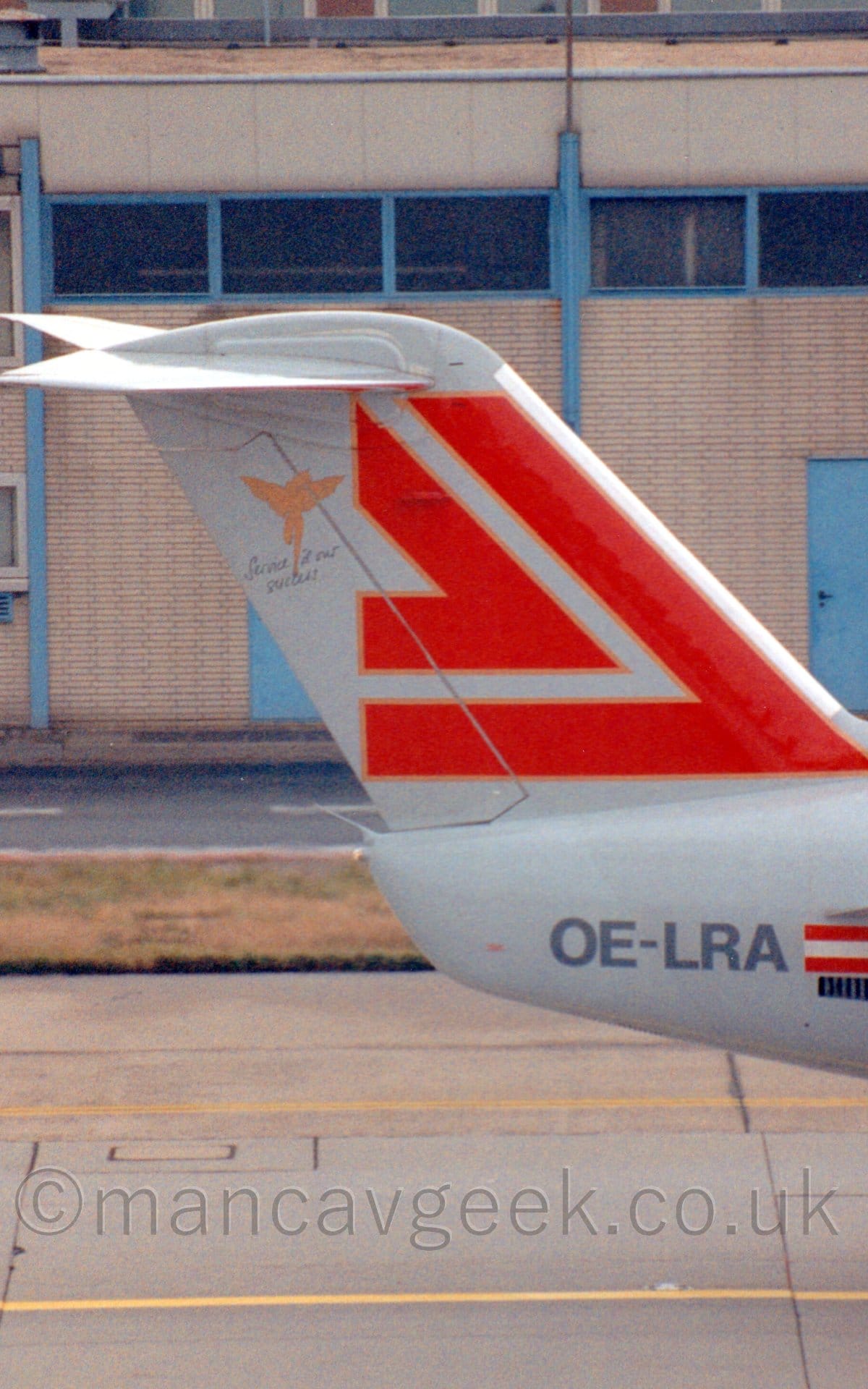 Close-up of the tail of a twin engined jet airliner taxiing from left to right. The plane is mostly white, with the grey registration "OE-LRA" nest to a small Austrian flag on the tailcone at the rear of the fuselage, and a pair of red chevrons outlined in gold and white on the white tail. A cream-coloured brick building with blue-framed windows fills the background.