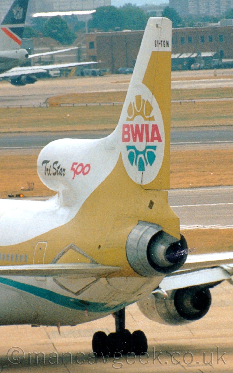 Vertically-oriented close-up of the tail of a 3 engined jet airliner taxiing from right to left. The plane is mostly white, with a yellowy-gold stripe running along the body, covering the passenger cabin windows, getting wider as it sweeps up into the tail. The front edge of the tail, and the intake for the centre engine at it's base, are white, with black text "TriStar" and the red number "500" on the engine intake. A white circle on the tail with red text "BWIA" in the middle, with 2 halves of a stylised steel drum, in the same yellowy-gold on top and in aquamarine below. The black registration "9Y-TGN" is at the top of the tail. Wide strips of grass line taxiways and a runway in the background, with the grey and dark blue tail of another jet airliner in the distance in the top left corner, with a brown building stretching across the frame from the right.