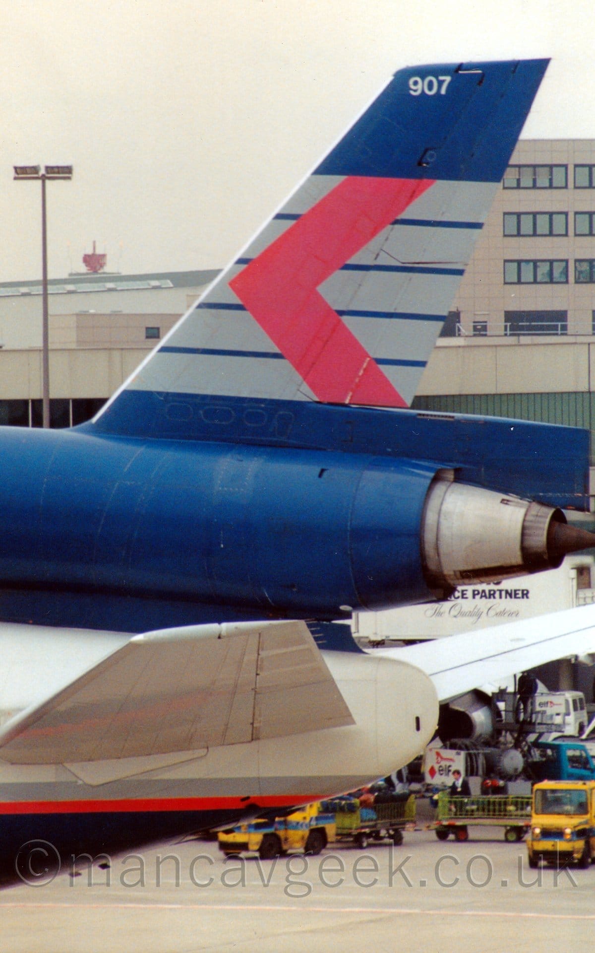 Close-up of the tail of a 3 engined jet airliner taxiing from right to left. The plane is mostly grey, with a dark blue belly, and a dark grey and red pinstripe separating the 2 colours. The tail and centre engine are dark blue, with 5 grey stripes running along the sides, overlaid with a red chevron pointing to the left, and the white number "907" at the top Yellow baggage trucks with blue body stripes can be seen in the background on the bottom right of the frame, with blue and white trucks behind them , while a grey terminal building can be seen over the top of this plane, under grey sky.