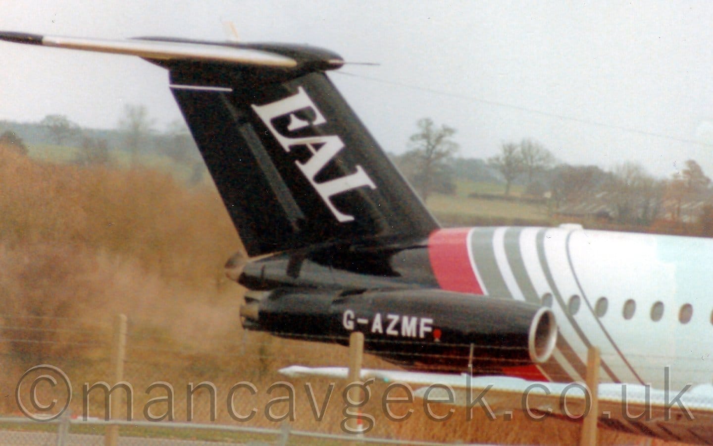 Close-up of the rear fuselage and tail of a twin engined jet airliner facing to the right, with the engines mounted on the sides of the rear fuselage. The plane is mostly white, with the rear fuselage being black, separated from the white section by a thick red diagonal band around the body, as well as 4 thinner grey bands. The tail and engine pods are also black. There are large white letters "EAL" on the tail, and the white registration "G-AZMF" on the sides of the engine pods. Several lengths of barbed wire, supported on concrete poles, stretch across the bottom of the frame in the foreground, with yellowy-brown brush and bushes on a slight incline in the background on the left. Green fields outlined by bushes and trees in the distance on the right slowly vanish into hazy grey sky.