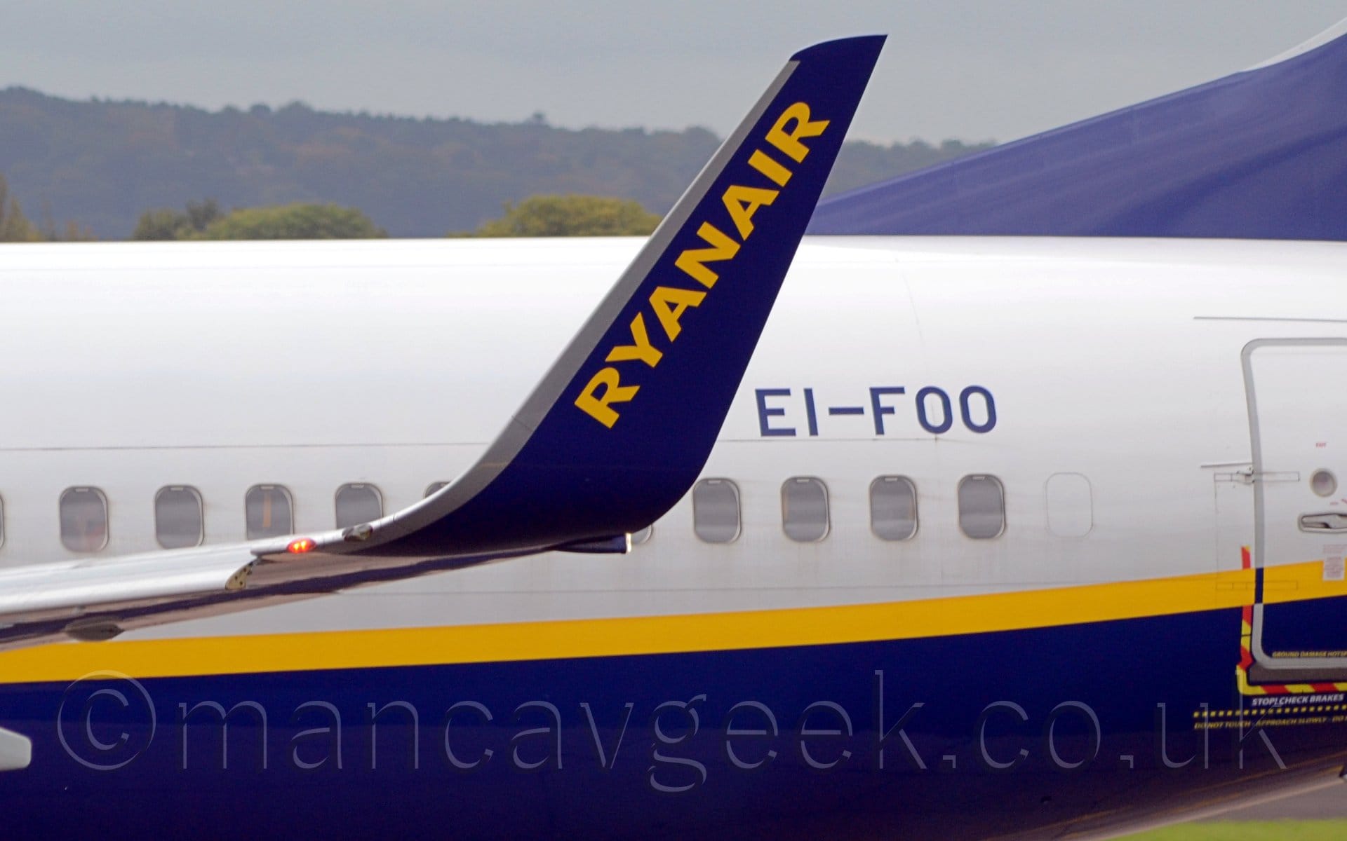 Close-up of the blue upturned wing-tip of a jet airliner taxiing from right to left. The plane is white on top with a blue belly, and a yellow stripe separating the 2 sections. There is diagonal yellow text "RyanAir" on the winglet, and blue registration "EI-FOO" on the upper rear fuselage. Tree-covered hills in the background lead up to hazy grey sky.