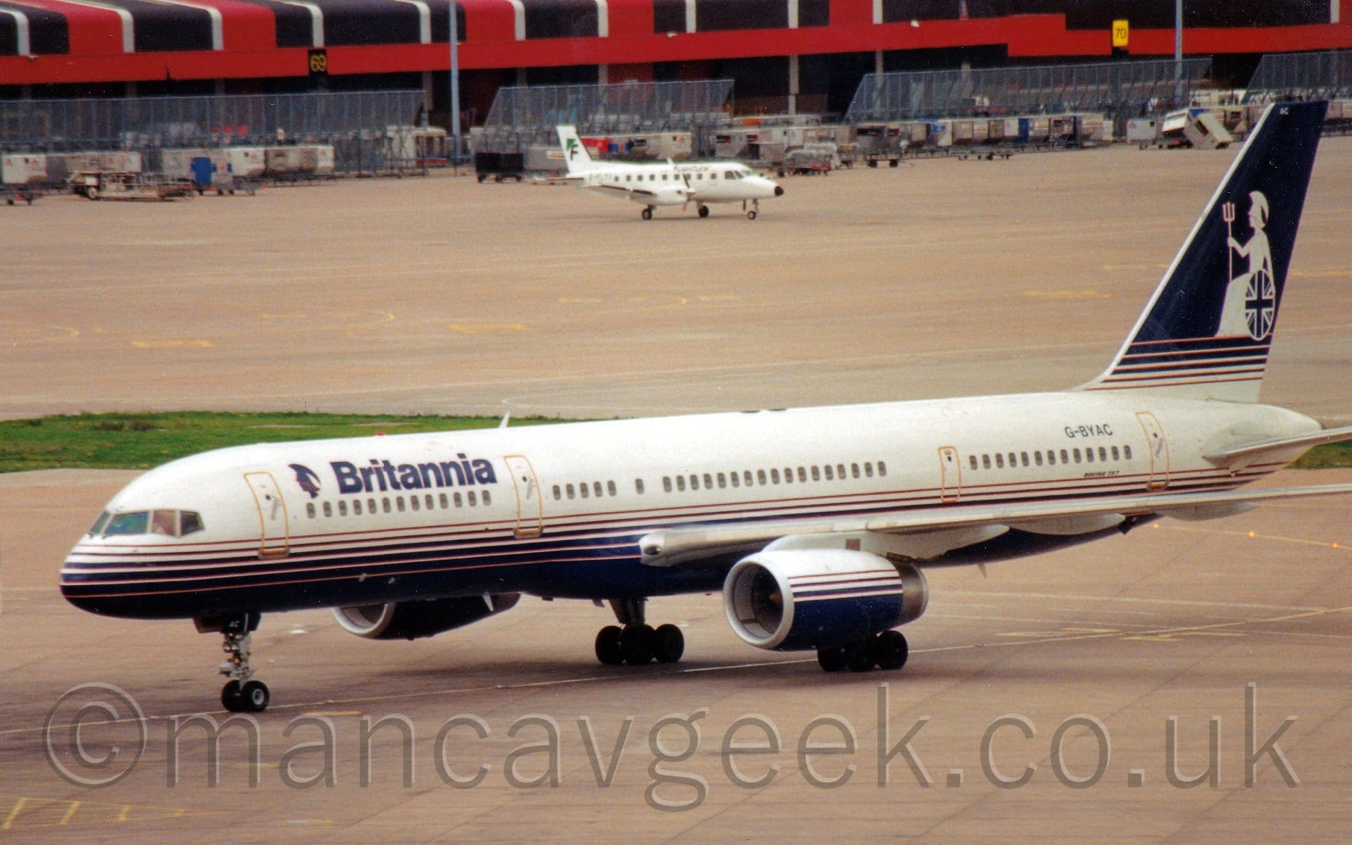 Side view of a twin engined jet airliner taxiing from right to left. The plane is mostly white, with a series of ever larger dark blue stripes running along the lower fuselage and belly. There are dark blue "Britannia" titles on the upper forward fuselage, behind a dark blue silhouette of a head wearing a plumed helmet. The black registration "G-BYAC" is on the upper rear fuselage, just below the tail. The tail itself has a white base, with dark blue stripes working up to a blue section covering the top 2/3rds. This blue section is overlaid with the seated image of a woman wearing an extravagantly plumed helmet, holding a large trident vertically in her right hand, and a circular shield with a variant of the UK flag in her left hand. Grey concrete apron fills the foreground, with more in the background, leading up to a red, brown, and white building at the top of the frame, with metal blast barriers and luggage containers lined up in front of it, along with a white, twin propellor-engined airliner.