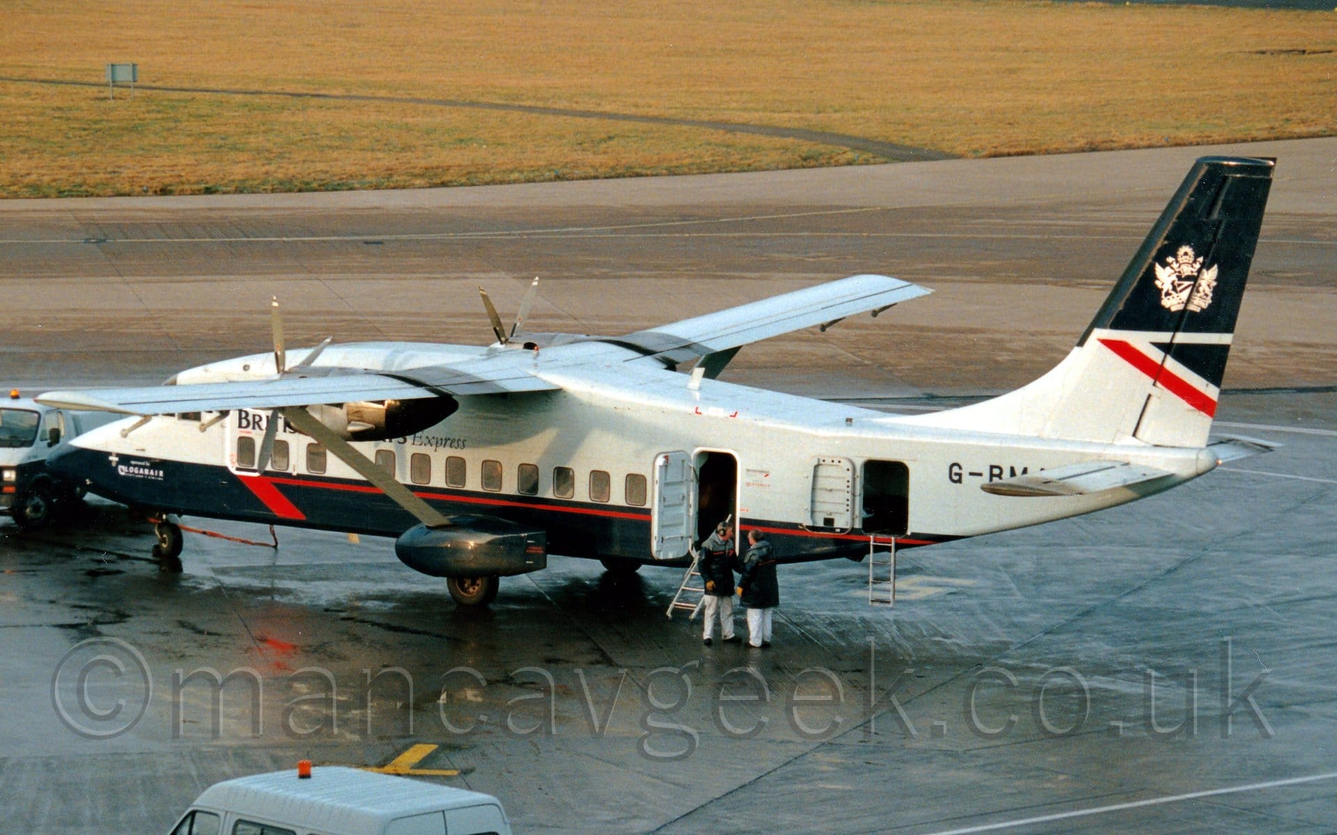 Rear side view of a high-winged, twin propellor-engined airliner parked facing top the left. The plane is largely grey, with a very dark blue belly, and a red stripe running along the lower body. Dark blue sponsons cover the fixed rear landing gear, connected to the bottom of the fuselage, with grey struts stretching out to support the long, thin wings just outboard of the engines. There are black "British Airways Express" titles on the upper forward fuselage, partially obscured by the engine pods under the wings, and the black letters "G-BM", part of the registration, on the upper rear fuselage. The tail is dark blue on the top and grey at the bottom, with a dark blue triangle and a diagonal red stripe in the middle, and a white Royal crest overlaid on the blue bit at the top. Slightly damp, black apron fills the foreground, with a the roof of a grey van with a red light on top visible at the bottom of the frame, and two men in dark blue and grey coats talking next to a pair of open doors in the rear fuselage. Gray taxiways in the background lead up to a large grassed area at the top of the frame.
