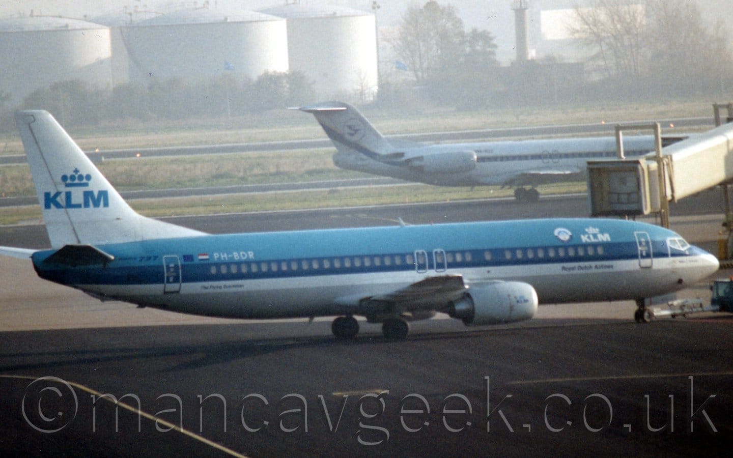 Side view of a twin engined jet airliner facing to the right but being pushed back to the left by a low-slung blue tug attached to the nose-wheel by a long white pole. The plane is light blue on top and grey on the bottom, with a thick dark blue stripe running along the body covering the passenger cabin windows, with a thinner white stripe just below that. There are white "KLM" titles under a stylised white crown on the upper forward fuselage, with a smaller, blue version of that logo on the white engine pods under the wings, and a larger, blue version on the white tail. A grey airbridge is hanging just behind this plane, with a white jet airliner with a pair of engines mounted on the rear fuselage is taxiing from left to right just beyond that. Black apron fills the foreground, marked with yellow lines for planes to follow. Black taxiways lined with green grass lead up large white fuel tanks in the distance, slowly vanishing into haze.
