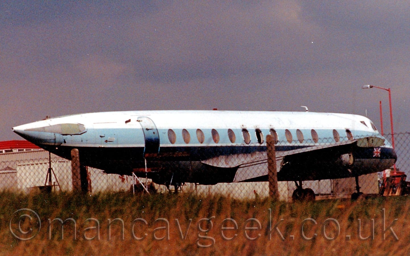 Rear side view of a 4 propellor-engined airliner parked facing to the right and slightly away from the camera. The plane is largely white, with a thick pale blue stripe running along the cabin covering the windows, a thick black stripe below that, and a grey belly. The engines and tail have been removed, as well as part of the wings and some of the cabin windows. A British flag is on the nose, just below the cockpit windows. In the foreground, long green and yellowy-brown grass leads up to a black chain-link fence on concrete supports, at the top of a slight rise. In the background, grey buildings with red roofs stretch across the frame from the left, under blue-grey skies.