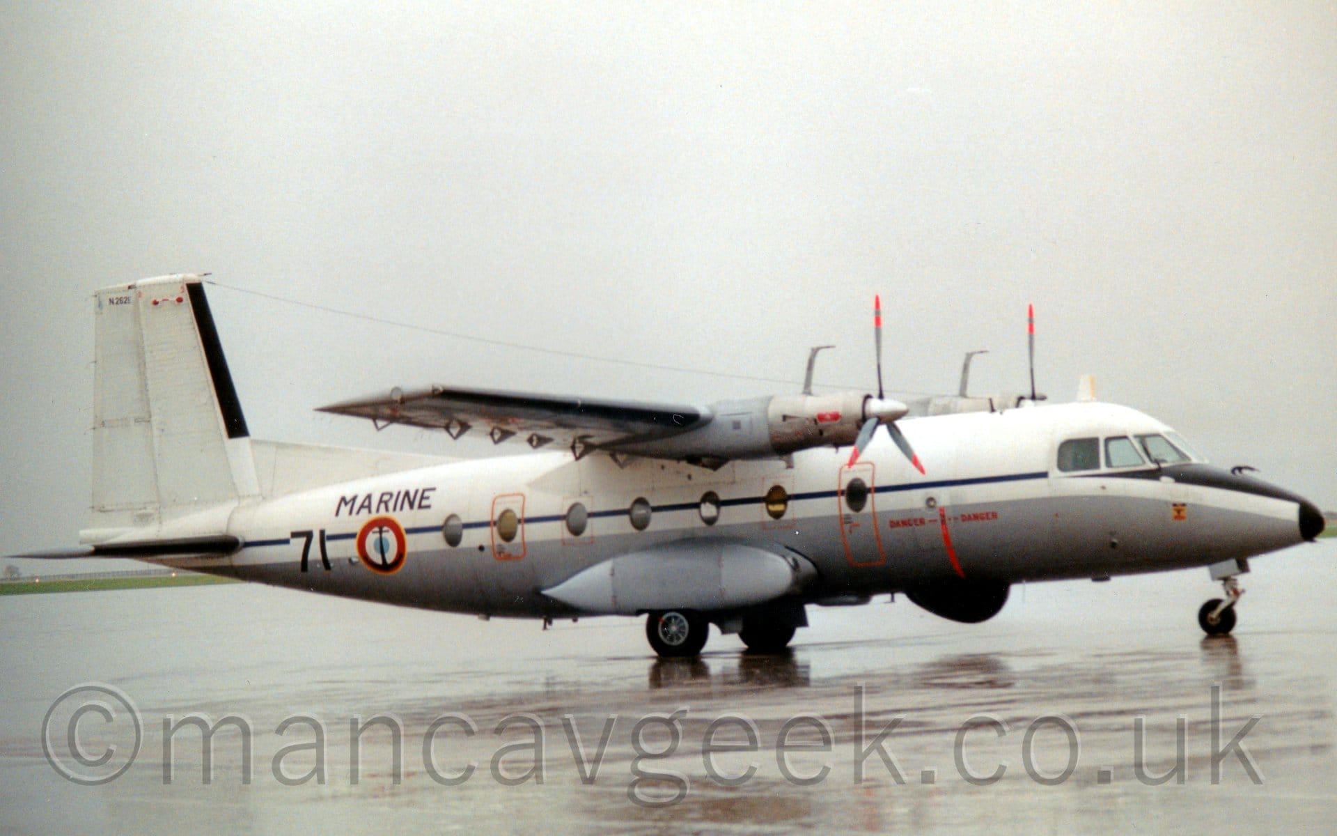 Side view of a high-winged, twin propellor-engined military transport plane parked facing to the right on a rather damp concrete apron. The plane is largely white, with a grey lower fuselage, and a black stripe running along the body, partially covering the cabin windows. There is a red, white, and pale blue roundel outlined in yellow on the rear fuselage, just ahead of the black number "71", under the black word "Marine". Grey concrete apron fills the foreground, under flat grey sky.