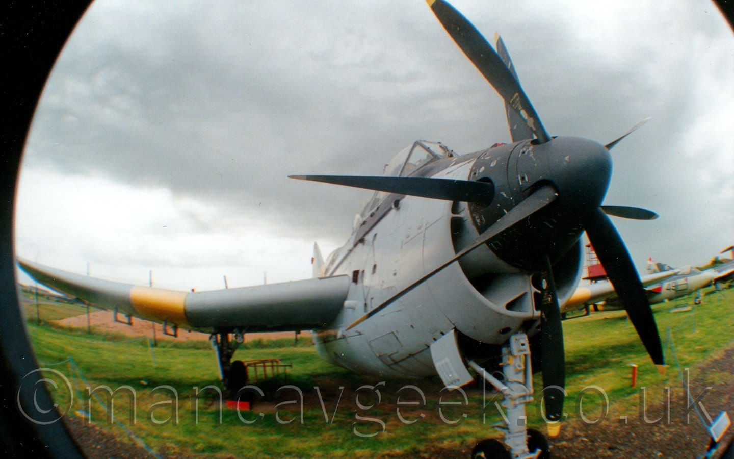 Front view of a twin propellor-engined, short and stubby military aircraft on display parked on concrete pads in the middle of a large grassed area, taken with a semi-fish-eye converter on a wide-angled lens, a combination that distorts straight lines while allowing a greater field of view at a closer range.. The 2 engines drive a set of contra-rotating propellors on the nose, each driving a different propellor on the same shaft, which explains the 2 sets of propellor blades. The plane is largely a pale grey-blue, with black around the cockpit canopy and around the propellors in the nose. There are yellow bands around the wings, just outboard of the main landing gear. Green grass fills the bottom of the frame, with more military planes in the background on the right. Dramatic grey sky fills the top of the frame.