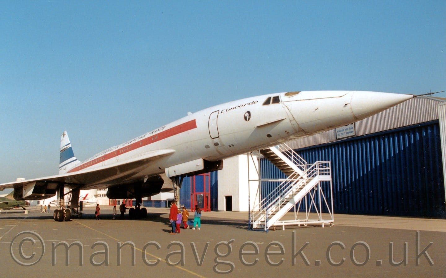 Side view of a long, thin, and extremely pointy, 4 engined jet airliner on display outside a museum, facing to the right. The plane is mostly white, with a red stripe running along the body, and a thick blue stripe bordered by thin blue stripes on the tail. There are small, black "Concorde" titles between the cockpit windows and the forward cabin door. The engines are mounted in pairs in square boxes under the wings. A set of tall, white, mobile air stairs is next to the forward door on the other side. A small family group in vividly coloured jackets and trousers is wandering around by the nose-wheel leg. A series of joined, white hangars with blue doors stretch across the background, with blue sky filling the rest of the frame.