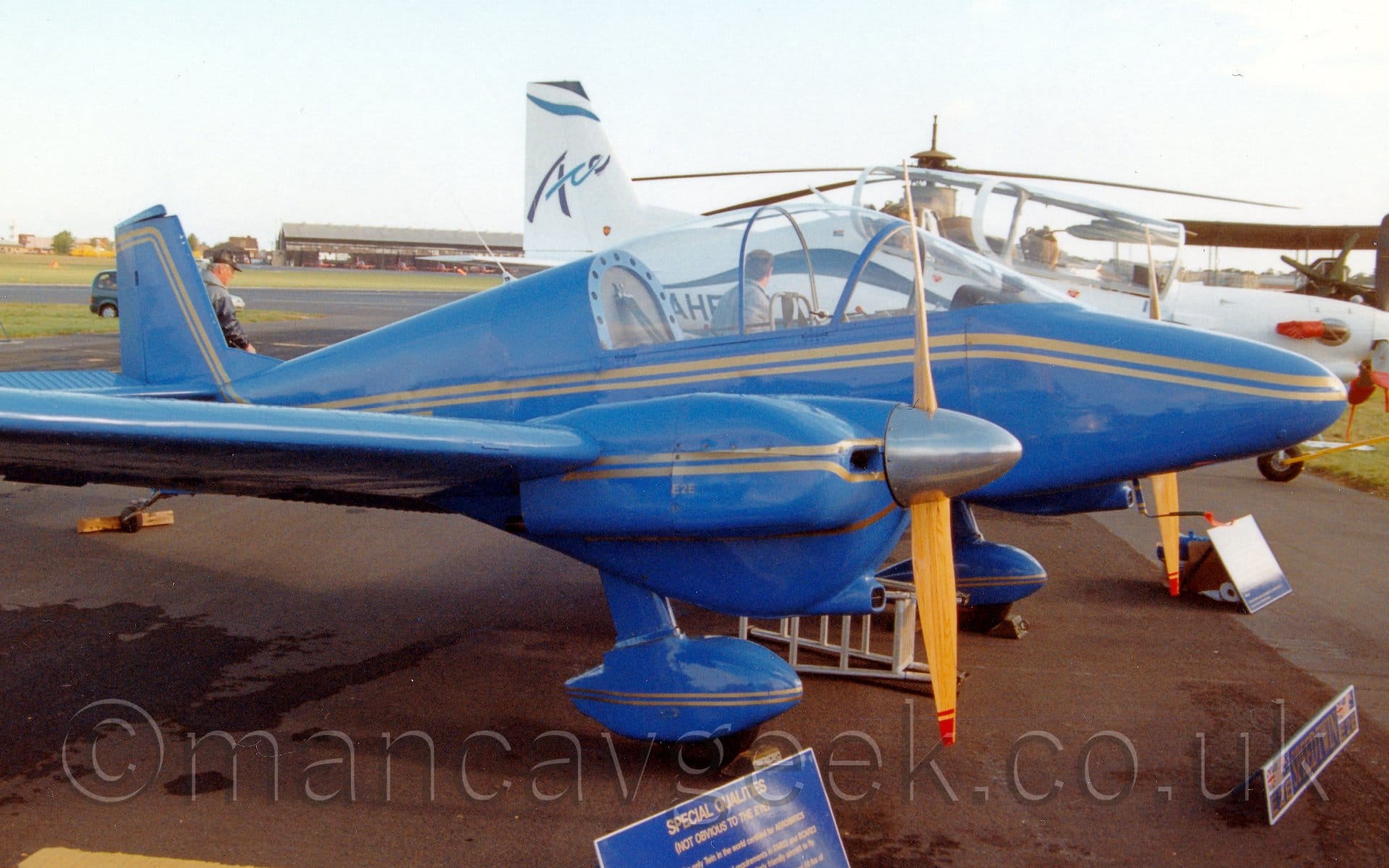 Side view of a twin propellor-engined light aircraft parked on tarmac facing to the right. The plane is a almost entirely blue, with a double gold stripe running along the body from the nose, as well as along the engine cowlings, just below the wings, and on the spats over the fixed undercarriage, suspended below the engine cowlings. A large bubble canopy covers the tandem 2-seat cockpit with a person wearing a grey jacket visible through the canopy, standing by the white plane on the far side. A military green helicopter and a large, twin engined bi-plane are visible in the background on the right, with a black hangar across the far side of the runway on the left, all under hazy grey sky.