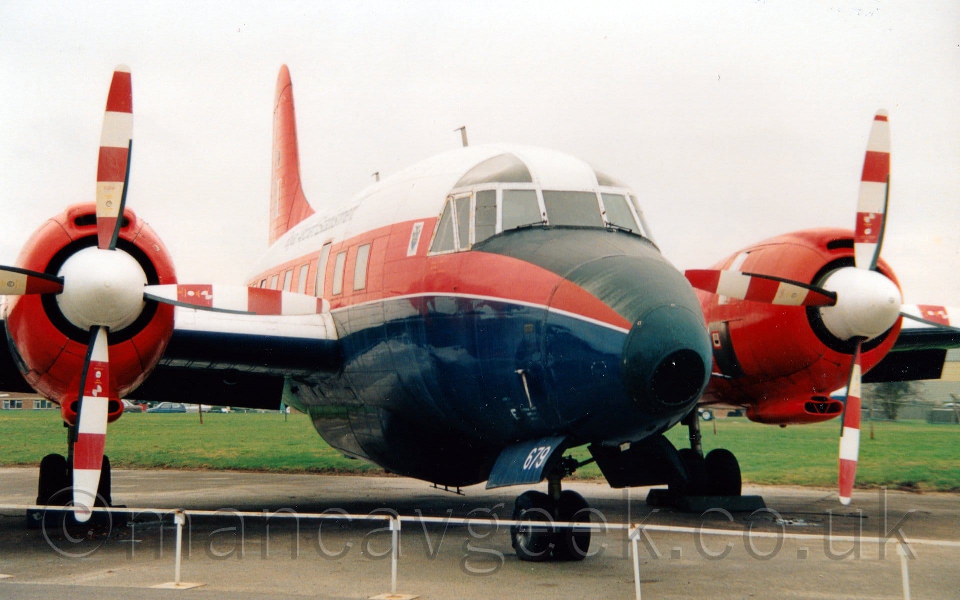 Front-side view of a twin propellor-engined military transport trainer aircraft parked facing to the right on tarmac behind a low, white, metal fence. The plane has a large red stripe running along the middle of the body, covering the passenger cabin windows, with a red roof and dark blue belly,. The engines mounted on the wing are the same bright red, as is the tail. The nose-cone, and top of the nose in front of the cockpit, is painted matt black. Green grass can be seen in the background, under the plane, with flat grey sky filling the rest of the frame.