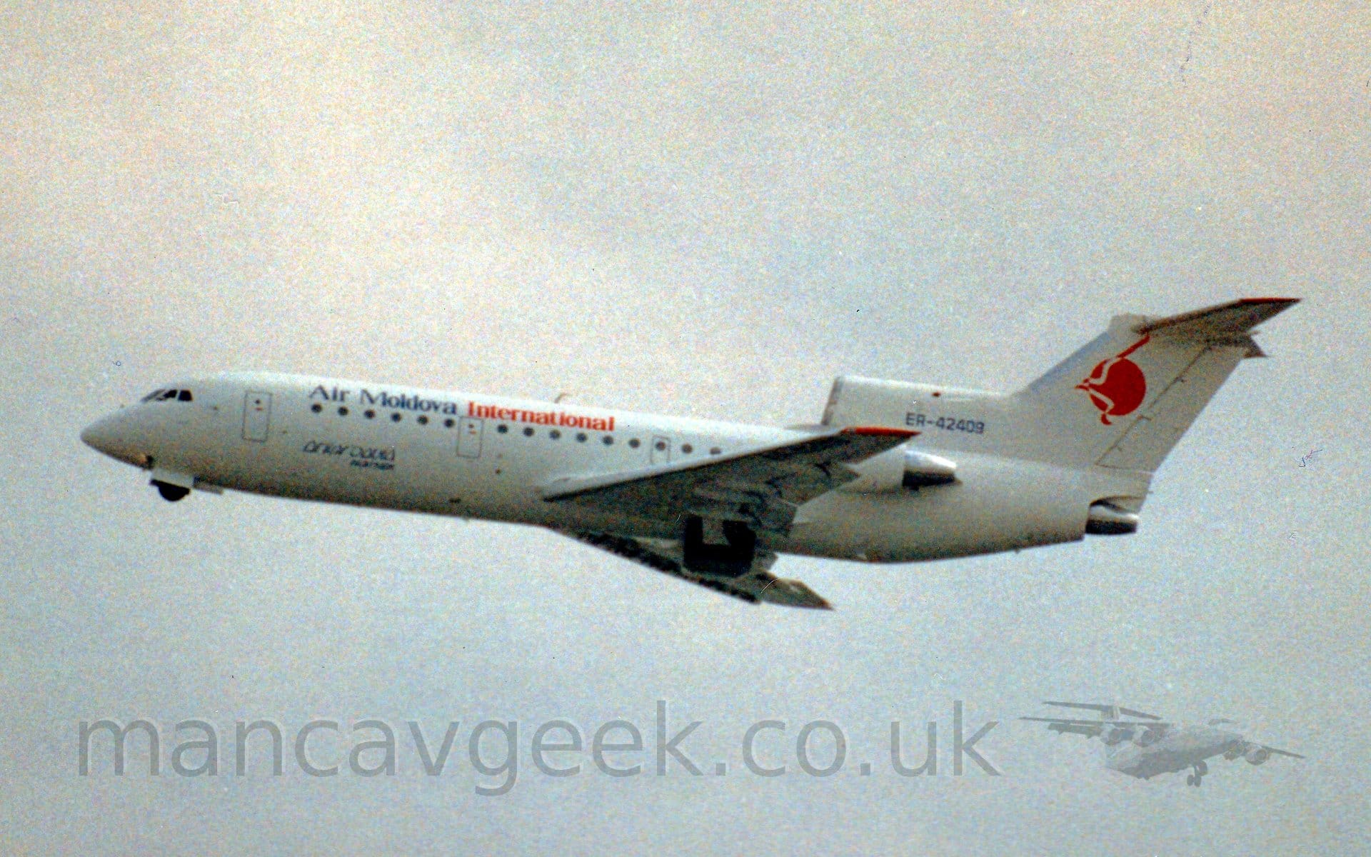 Somewhat grainy side view of a 3 engined jet airliner flying from right to left, with flaps extended behind the wings, the undercarriage being retracted, and the nose raised sharply, suggesting it has just taken off. The plane is largely white, with dark blue "Air Moldova" and red "International" titles on the upper forward fuselage, and the black registration "ER-42409" on the centre engine intake in front of the tail. A red circle adorned with a red and white stylised flying bird adorns the tail. The white engine pods on the rear fuselage are partially hidden by the wing. Grey sky fills the rest of the frame.