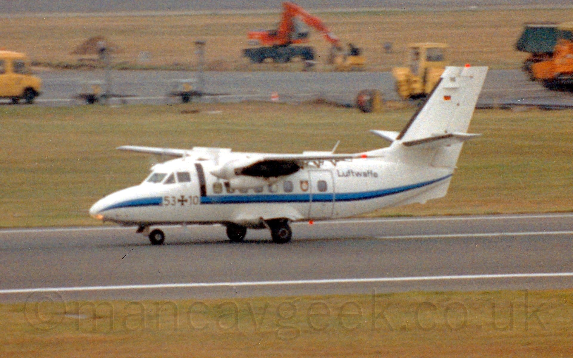 Side view of a high-winged, twin propellor-engined military transport aircraft moving from right to left along a black runway, with flaps extended from the rear of the wings, and spoilers deployed from the top of the rear of the wings, suggesting it has just landed and is slowing down. The plane is largely white, with a light and dark blue stripe running along the body, overlaid with the number "53+10" under the cockpit windows. There are black "Luftwaffe" titles on the rear fuselage. Green grass lines the runway in the foreground, with more grass in the background, with various yellow or orange construction vehicles on a black taxiway in the distance.