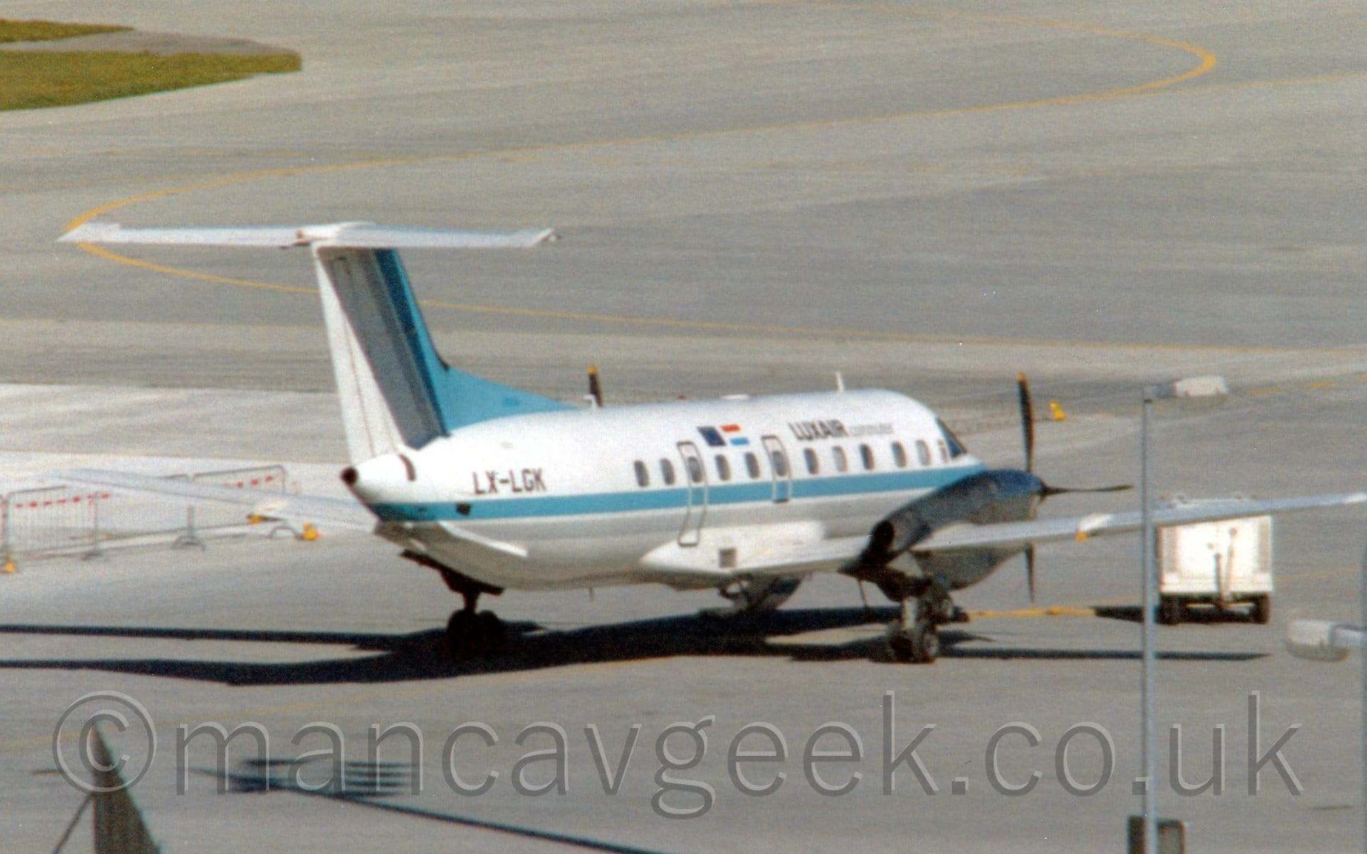 Rear side view of a twin propellor-engined airliner parked facing to the right and away from the camera. The plane is mostly blue, white, with a grey belly, and a blue stripe running along the body. The tail is mostly white, with the leading edge being pale blue, with a thin grey stripe. There are black "LuxAir" titles on the upper forward fuselage, along with the black registration "LX-LGK" on the upper rear. Grey concrete apron fills the foreground and most of the background, with a tall light pole coming in to view in the bottom right corner, and a patch of grass in the top left.
