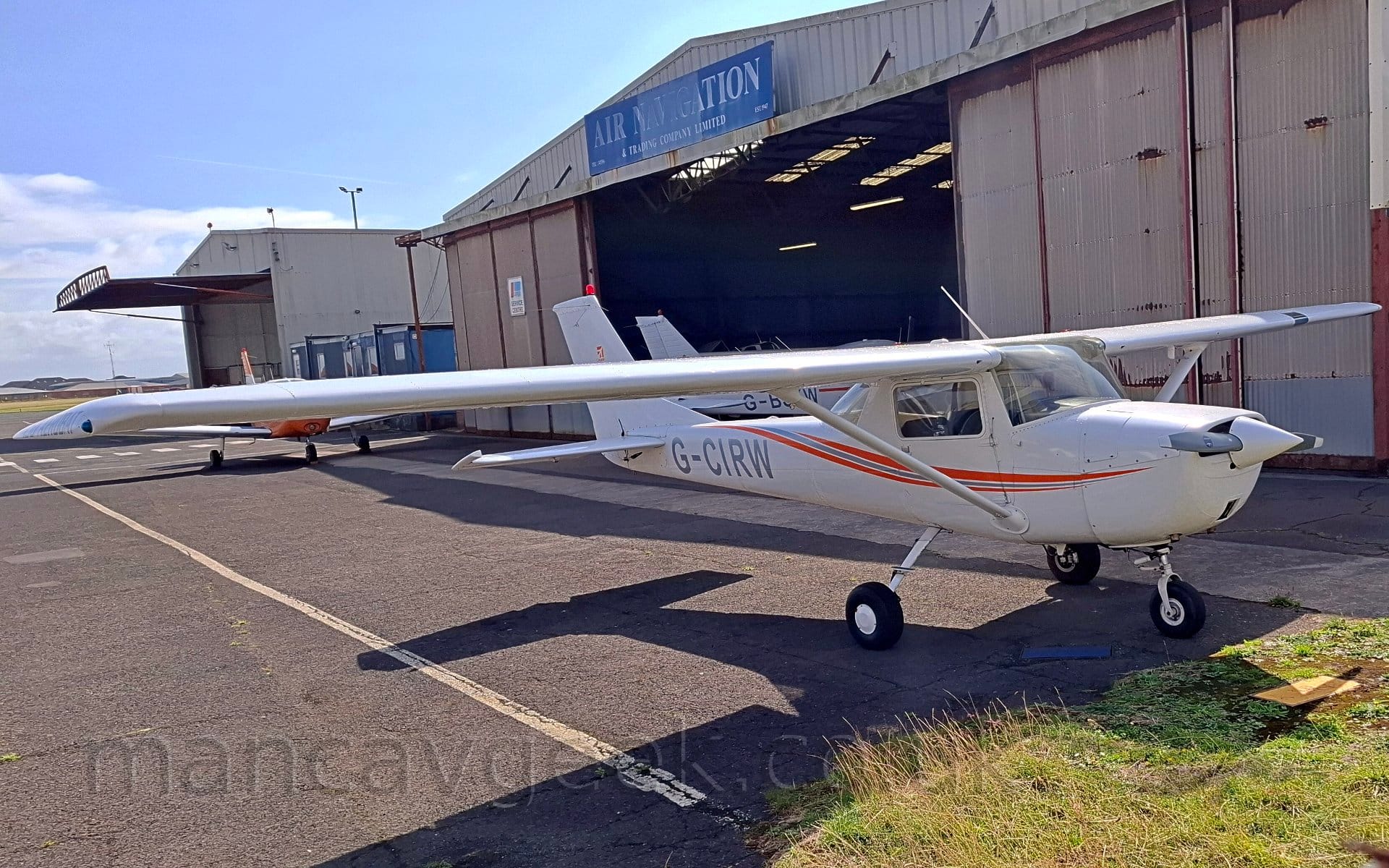 Side view of a single engined, high-winged light aircraft parked facing to the right. The plane is white, with an orange and grey stripe running along the body from the propellor, sweeping up the top just behind the cockpit. The grey registration "G-CIRW" is on the rear fuselage. Black tarmac apron fills most of the oreground, with a patch of grass in the bottom right corner, directly in front of the plane. A pair of old grey hangars dominate the background, the nearest one with it's doors wide open, giving a hint of it's inhabitants. Blue sky with fluffs of white cloud fills the rest of the frame.