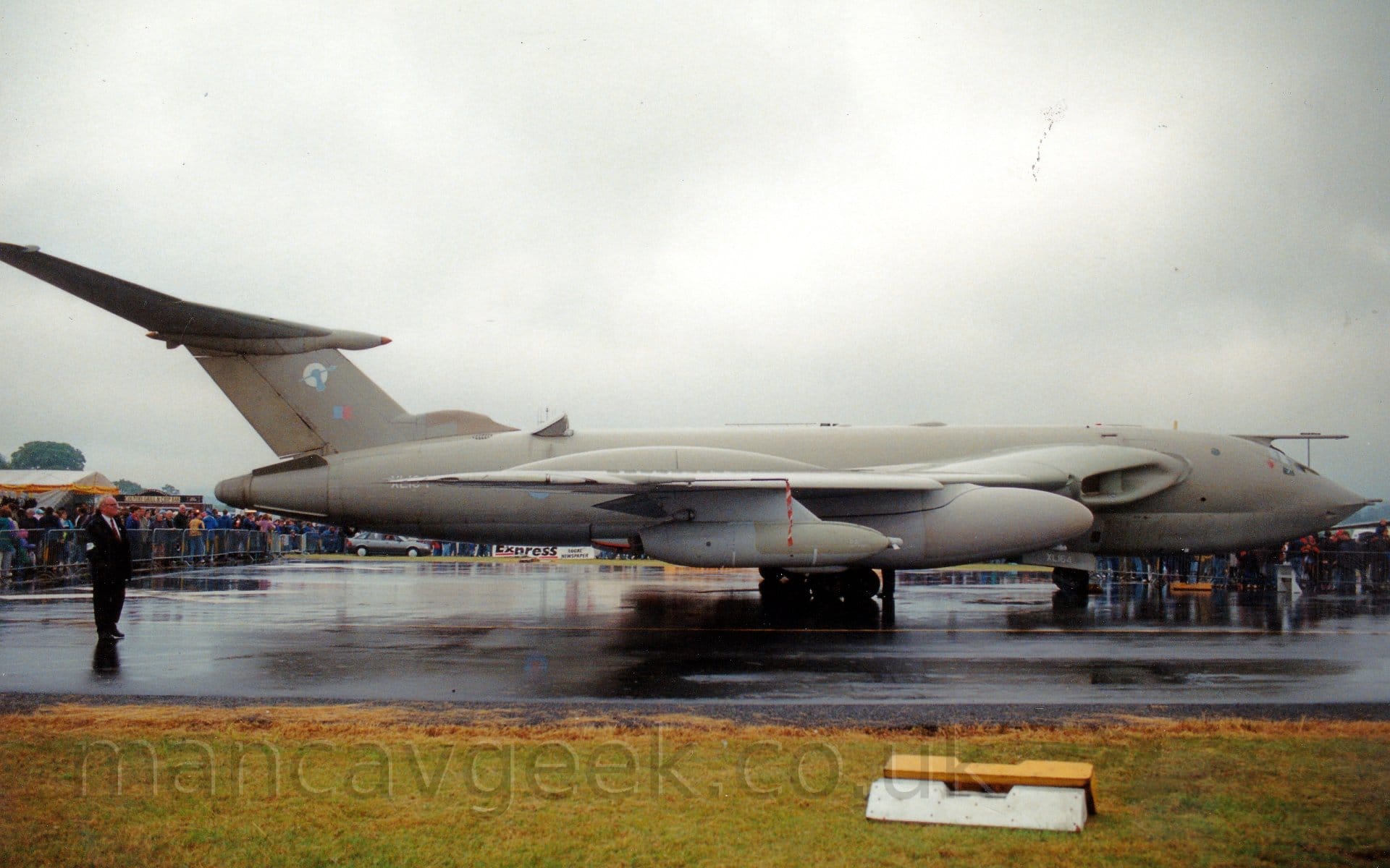 Side view of a rather bizarre-looking, 4 jet engined military tanker aircraft parked facing to the right on a black taxiway. The plane is painted mostly in a dark , sandy grey-brown colour scheme on the upper surfaces, with the belly a slightly lighter shade. The plane looks rather 1950's style "retro-futuristic", a sharp pointed nose with sleek flowing lines along the body, leading to the curved rear fuselage. The engines are buried in the wing, with large intakes in the wing root, with the wings curving out slightly to the side, with large pods under the wing containing large fuel tanks, and smaller pods out-board of those. The highly swept T-tail is mounted on the upper rear fuselage, hanging back well past the end of the body, with a pale blue and red fin flash, and a white circle overlaid with a blue arm holding a spear. Green grass lines the taxiway in the foreground, with crowds of people behind metal barriers clustered around the plane in the background, under dreary grey sky.
