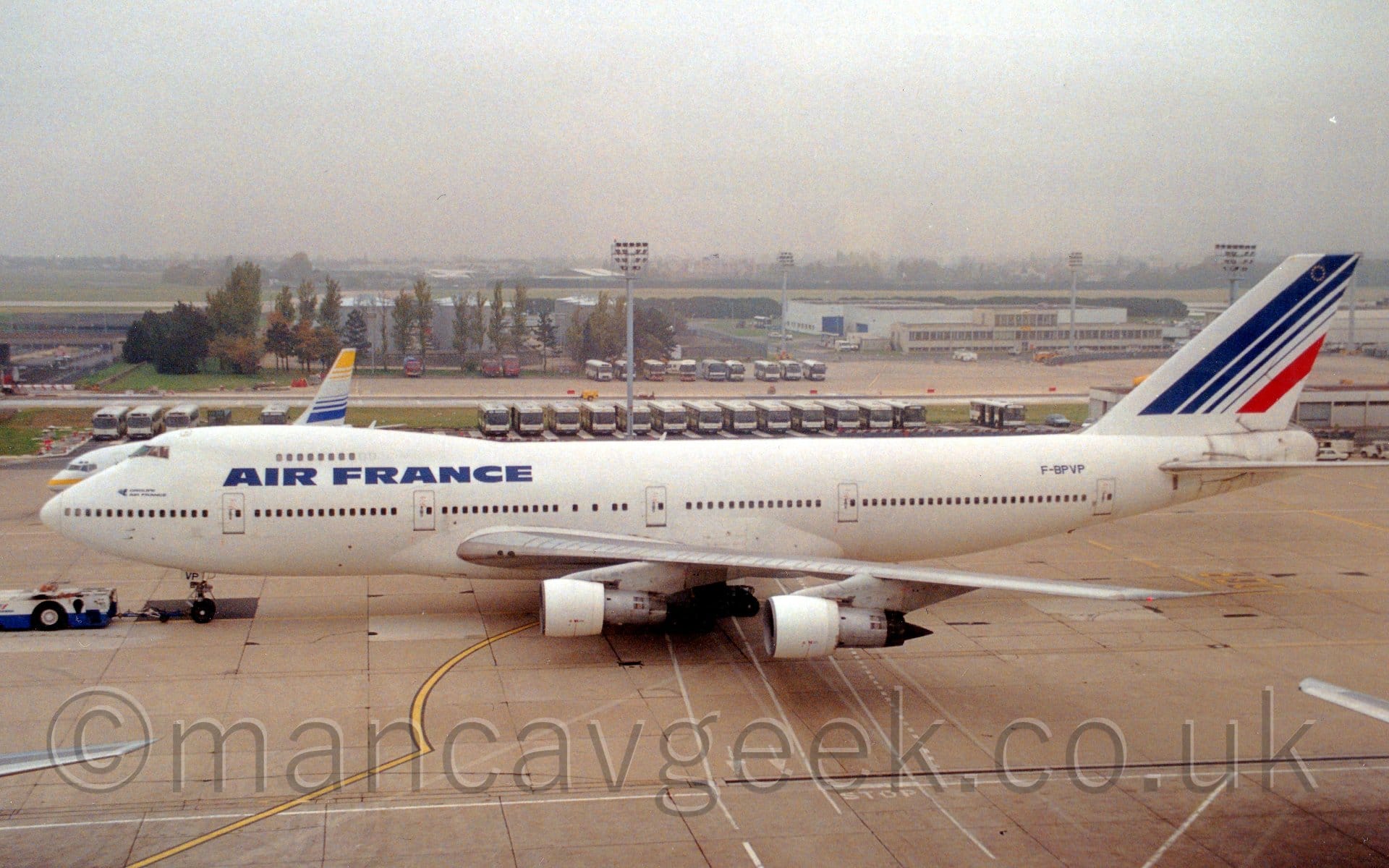 Side view of a very large, 4 engined jet airliner being towed from right to left by a low-slung white and blue tug attached to the nose-wheel by a long black pole. The plane is almost entirely white, with large, blue, "Air France" titles on the upper forward fuselage, between the 2 passenger decks. The blue registration "F-BPVP" is on the upper rear fuselage, and the last 2 letters are on the open nose-wheel doors. The tail is white, with a series of 4 diagonal blue lines, getting thinner as they move rearwards, and a final, thick red stripe at the rear. Grey concrete apron fills the foreground, with rows of buses parked side-by-side in the background, and white buildings and trees in the distance, slowly vanishing into the hazy grey sky.
