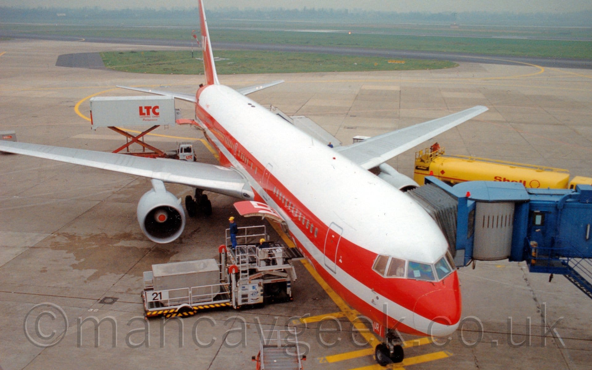 Almost head-on view of a twin engined jet airliner parked facing slightly to the right of the camera. The plane is largely white, with a red belly, and a thick red stripe running along the body, covering the passenger cabin windows. There are white "LTU Sud" titles on the lower forward fuselage, as well as on the red tail. The white, last 2 letters of the registration, "UR", are on the open nose-wheel doors. The plane is pretty much perfectly parked on a barred, yellow line on the concrete apron, with a blue and grey air bridge attached to the forward cabin door on the left side. There is a freight loader parked next to the open lower fuselage cargo door, with a container ready to be lifted. A yellow fuel tanker with red "Shell" titles is parked in front of the left wing, on the other side of the air bridge. A white catering truck with red "LTC" titles on the side is parked by the rear cabin doors, with it's cargo bed raised to allow access to the plane. In the background, green grass lines a runway, with more grass and trees in the distance, slowly vanishing in to haze.