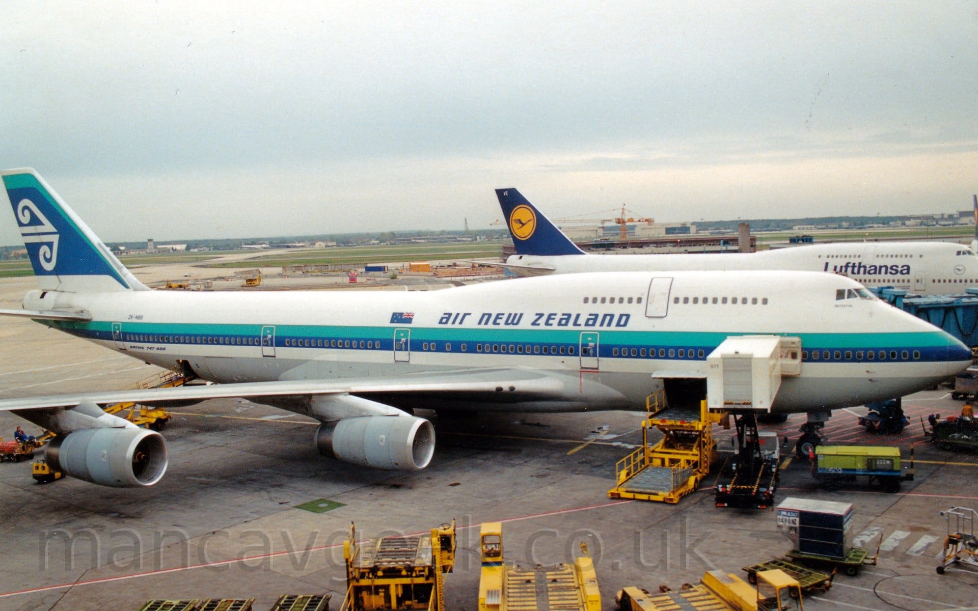 Side view of a very large, 4 engined jet airliner parked at an airport terminal, facing to the right. The plane is mostly white, with an aquamarine and blue stripe running along the body, the blue part covering the passenger windows on the main deck. There are blue "Air new Zealand" titles on the upper forward fuselage, next to a New Zealand flag. The tail is mostly blue, outlined with a thin aquamarine stripe on the top and white at the bottom, with a pair of stylised white waves in the middle. Yellow baggage loaders litter the grey concrete ramp in the foreground, some connected to open hatches in the lower fuselage. In the background, another very large jet airliner with a similar shape, but with a dark blue tail with a dark blue stylised flying bird in a yellow circle in the middle, with dark blue "Lufthansa" titles on the upper forward fuselage. Beyond that plane, a general view across the airfield sees extensive areas of green grass separated by taxiways and runways, with planes and buildings in the distance, under grey skies.