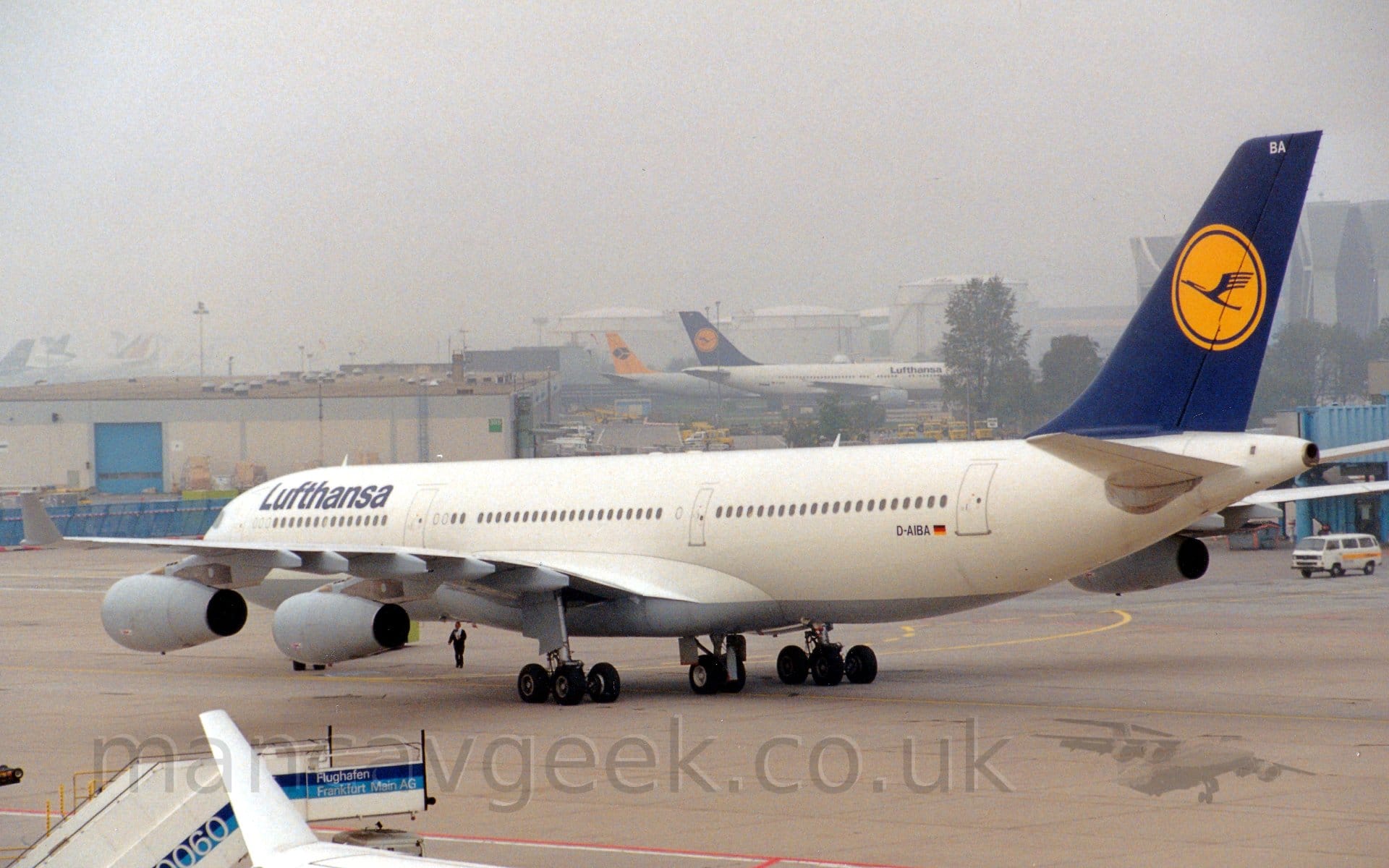 Rear side view of a 4 engined jet airliner taxiing from right to left, turning sharply to it's right. The plane is mostly white, with a grey belly, and dark blue "Lufthansa" titles on the upper forward fuselage. The tail is dark blue, with a yellow circle, with a stylised dark blue flying bird in the middle. Grey concrete apron fills the foreground, with the white winglet of a jet airliner next to a white and blue mobile set of airstairs visible at the bottom left of the frame. A low, grey building is on the left of the background, with some trees on the right. A pair of white jet airliners, one with a blue and yellow tail, the other yellow and black, are in the distance, in front of some large fuel storage tanks. Murky, misty grey sky fills the rest of the frame.
