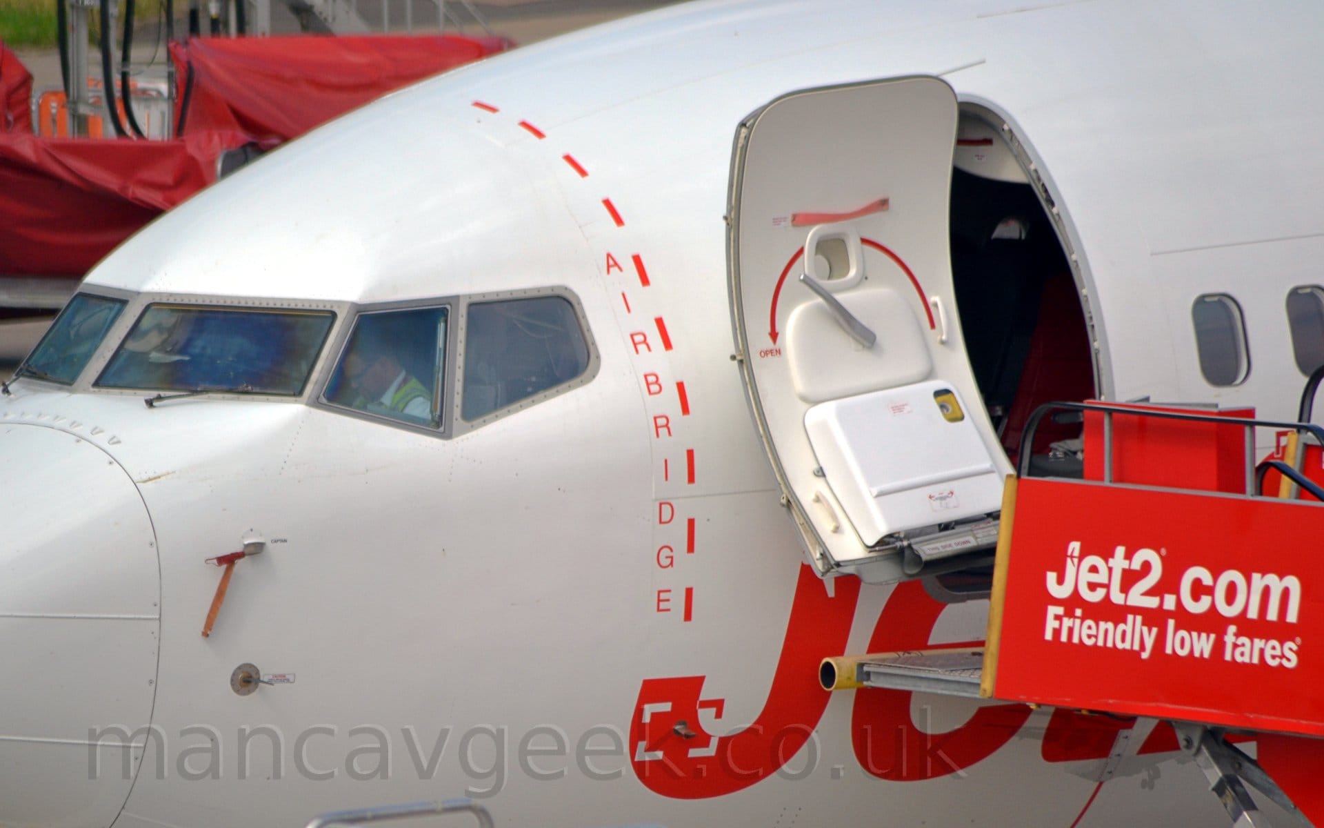 Close-up detail of a white jet airliner parked facing to the left showing the area around the open forward cabin door. A red set of mobile airstairs with white "Jet2.com" above "Friendly Lower Fares" titles has been positioned by the open door, a yellow bumper leaning against the side of the fuselage just below the door. Large, red "Jet2" titles are on the side of the plane just below the door, but are largely obscured by the airstairs. Between the door and the cockpit windows, there is a dashed red line with the word "Airbridge" arranged vertically on the left.