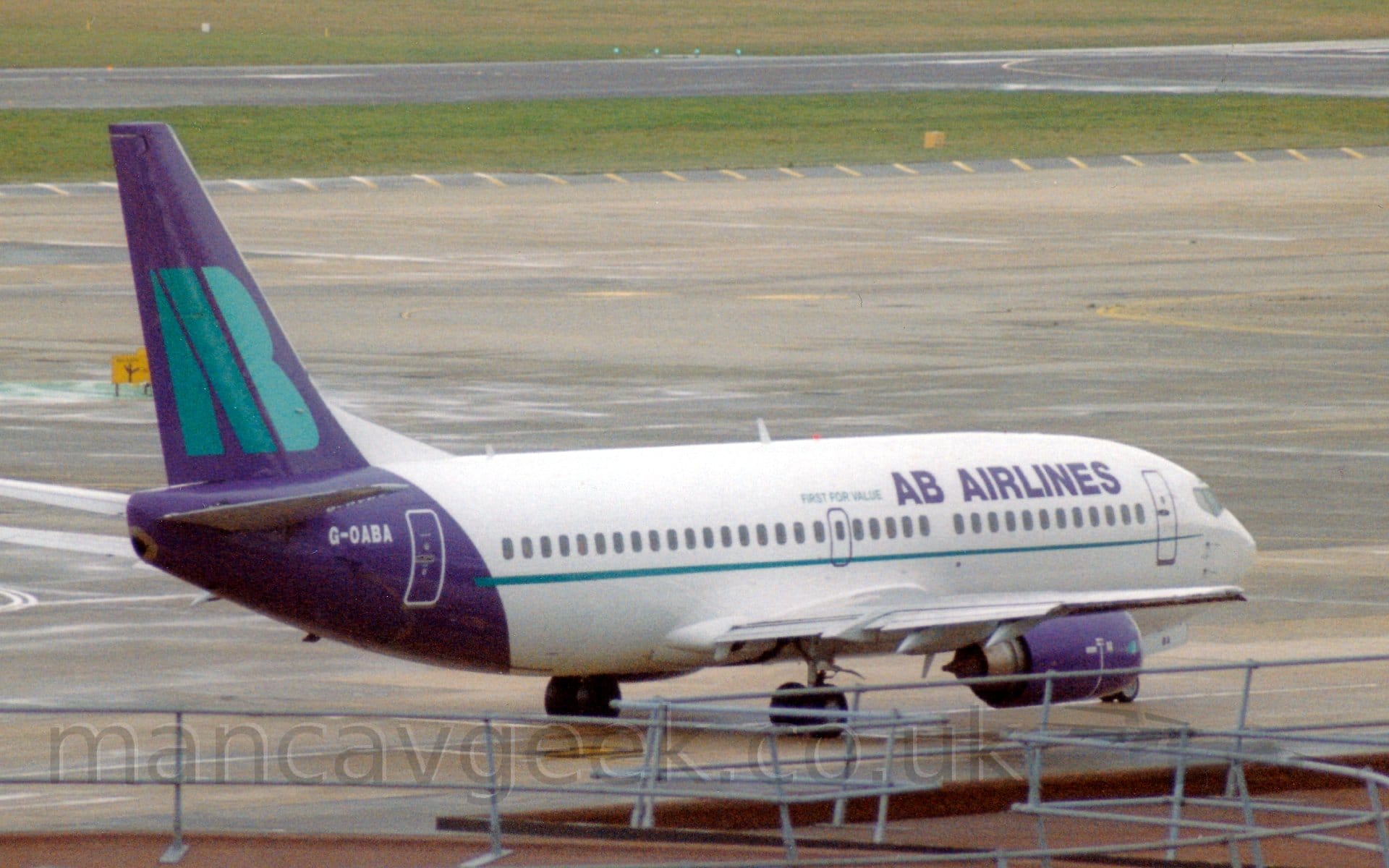 Side view of a twin engined jet airliner taxiing from left to right. The plane is mostly white, with purple "AB Airlines" on the upper forward fuselage, with smaller green "First For Value" text aft of that. A thin green stripe runs along the body, under the passenger cabin windows. The rear fuselage and tail is purple, with the white registration on top of the fuselage, and large, green, letters "AB" on the tail. The roof of a terminal building fills the foreground, with metal railings on top . Grey concrete apron fills the background, with green grass lining the sides of a black runway in the distance.