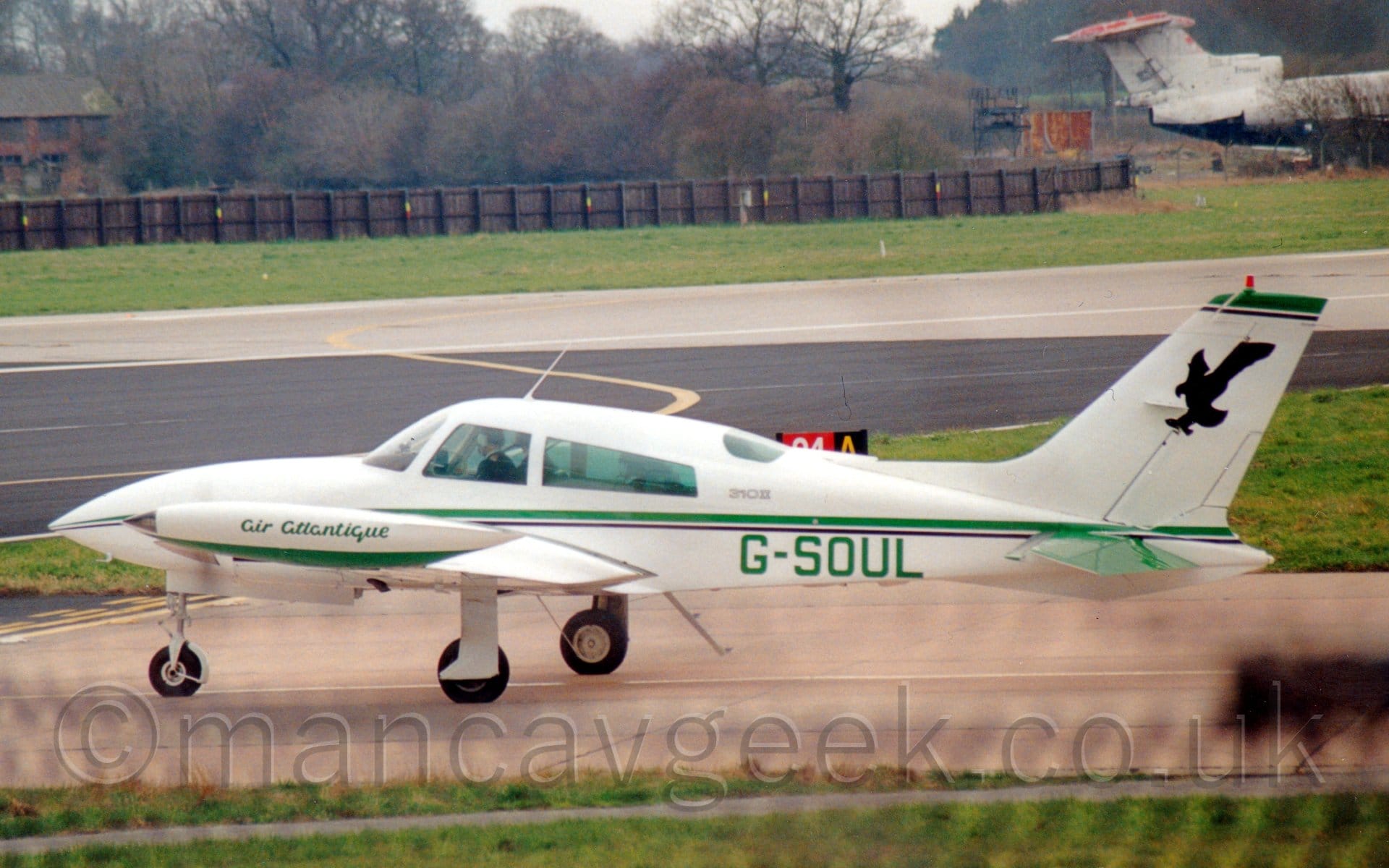 Side view of a twin engined light aircraft taxiing from right to left along a reddish-grey taxiway. The plane is mostly white, with a green and black stripe running along the body, with the green registration "G-SOUL" on the lower rear fuselage. There are green "Air Atlantique" titles on the wing-tip fuel tanks, and a black shadow of a stooping eagle on the tail. Green grass lines the taxiway in the foreground, with more grass lining both sides of the runway in the background. A black wooden fence stretches across the background from the left, with the tail and rear fuselage of a rather forlorn 3 engined jet airliner coming in to view of the right. Trees in the distance meet bright but hazy sky.