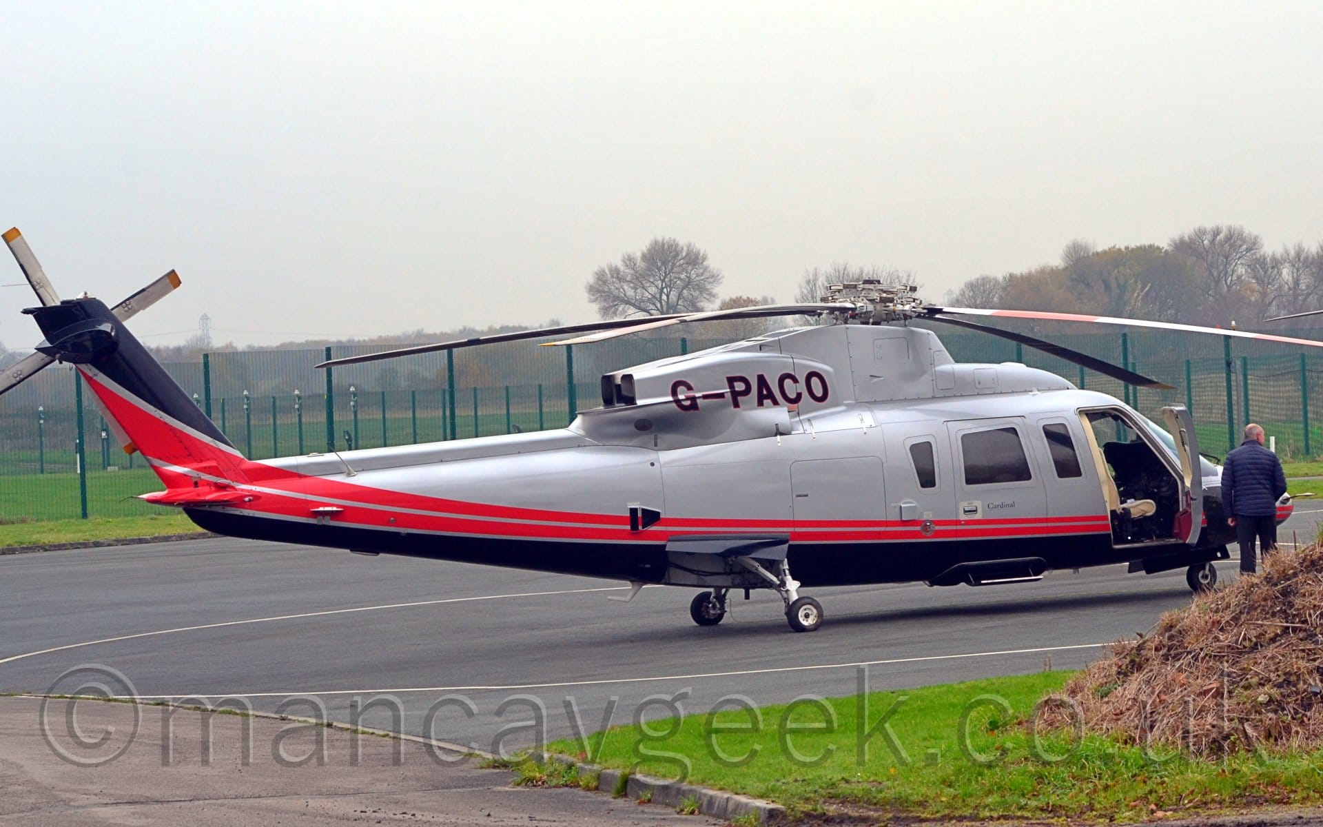 Side view of a helicopter parked facing to the right on black tarmac apron. The chopper is mostly grey, with a pair of red stripes running along the body and up in to the tail, and a black belly. The black registration "G-PACO" is on the rear of the engine cowling on the top of the airframe, outlined in red. The cockpit door is wide open, showing part of the cockpit, with a man in a blue jacket standing beside it.. There are small black "Cardinal" titles on the passenger door. In the foreground, there is a patch of lush green grass, with all sorts of grass clippings and similar item piled up in the corner. Tall green chainlink fences are in the background, with trees in the distance, under bright but hazy sky.
