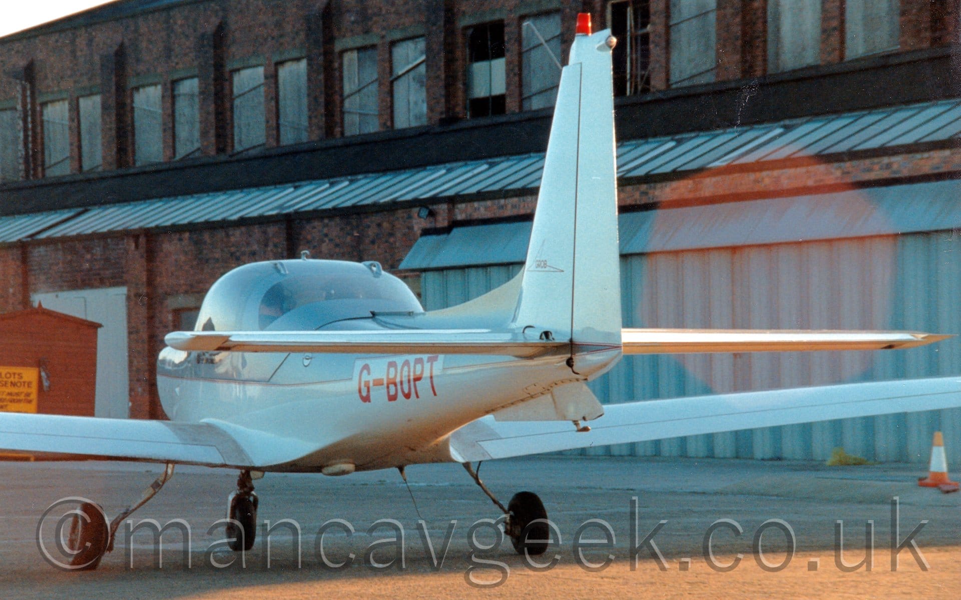 Rear view of a single engined light aircraft parked on tarmac facing to the left and away from the camera. The plane is grey, with a white lower half, outlined in red, with red registration "G-BOPT" on the rear fuselage. A large, red brick hangar fills the background, some of it's windows in the upper storey looking a bit sorry for themselves.