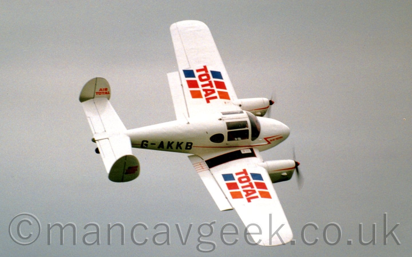 Side view of a twin engined light aircraft flying from left to right, highly banked to the right. The plane is mostly white, with a thin red stripe running along the body, overlaid with the black registration "G-AKKB" on the rear fuselage. There are red "Total" logo's on each wing, with diagonal blue, red, and orange stripes running through, and smaller red "Air Total" titles on both the inside and outside faces of the twin tails. Grey sky fills the rest of the frame.