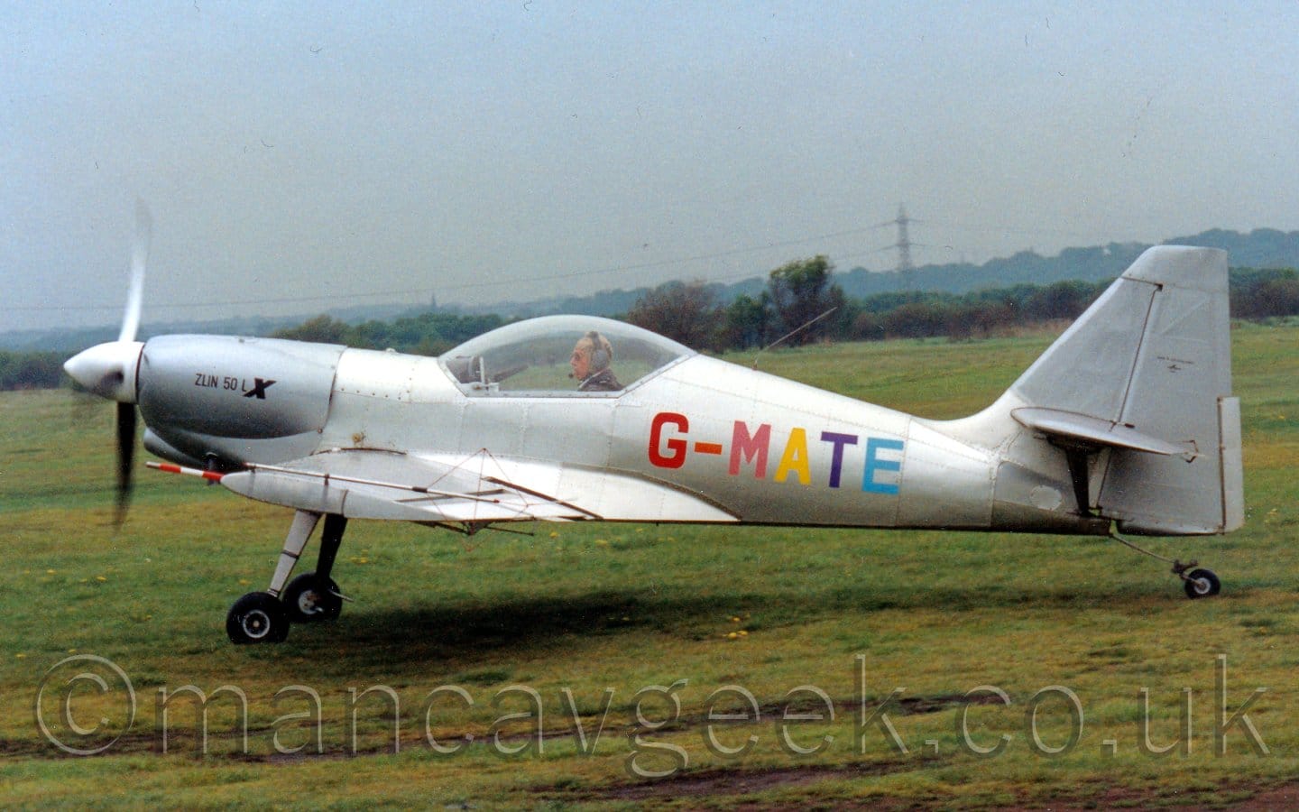 Side view of a single-engined light aircraft taxiing from right to left on a grass airfield. The plane is in a bare metal finish, with the multi-coloured registration "G--MATE" in red, salmon pink, yellow, purple, and light blue. The black text "Zlin Z50 LX" is on the engine cowling on the nose, the last letter being larger and bold. The head and shoulders of the pilot can be seen through the large bubble canopy, wearing a brown leather jacket over a white shirt, and a grey and black headset over a grey cloth flying helmet. Green grass fills the foreground and most of the background, with trees in the distance, under dismal grey sky.