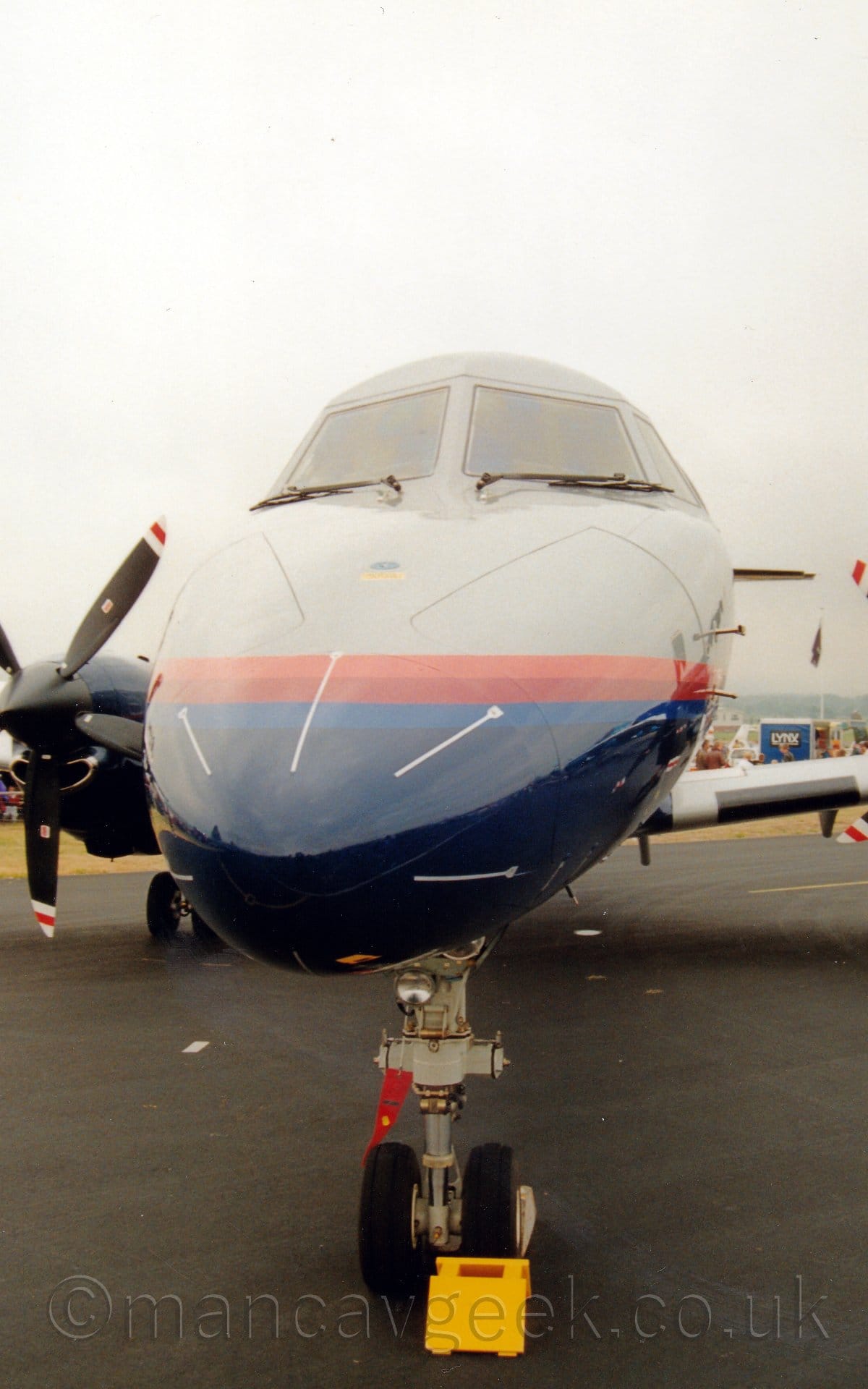Close-up front view of the nose of a twin propellor-engined airliner parked on black tarmac, more or less facing the camera. The plane is grey, with a dark blue belly, the 2 colours separated by an orange, red, and mid-blue stripe running along the body. The planes right-hand engine can be seen on the left of the frame. The nosewheel leg has a red tag attached to a pin just below the landing light, and there is a yellow chock in front of it, to prevent the plane from moving, with a white chock behind the wheel. In the background, part of the crowd can be seen, along with an advertising hoarding for Lynx deoderant. Grey sky fills the rest of the frame.