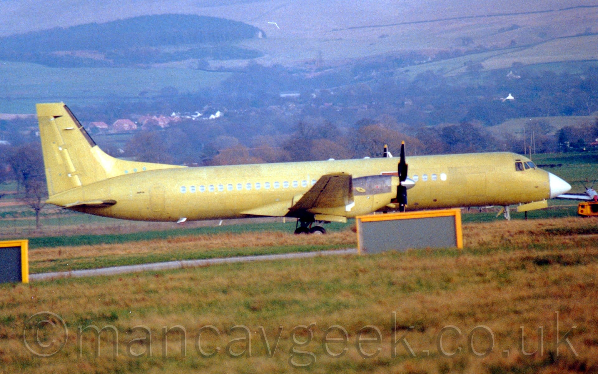 Side view of a twin propellor-engined airliner parked facing to the right. The plane has a white nose cone, with the rest covered in a yellowy-green primer. Brown grass with green patches fill the foreground, with rolling hills filled with fields, trees, and houses in the background.