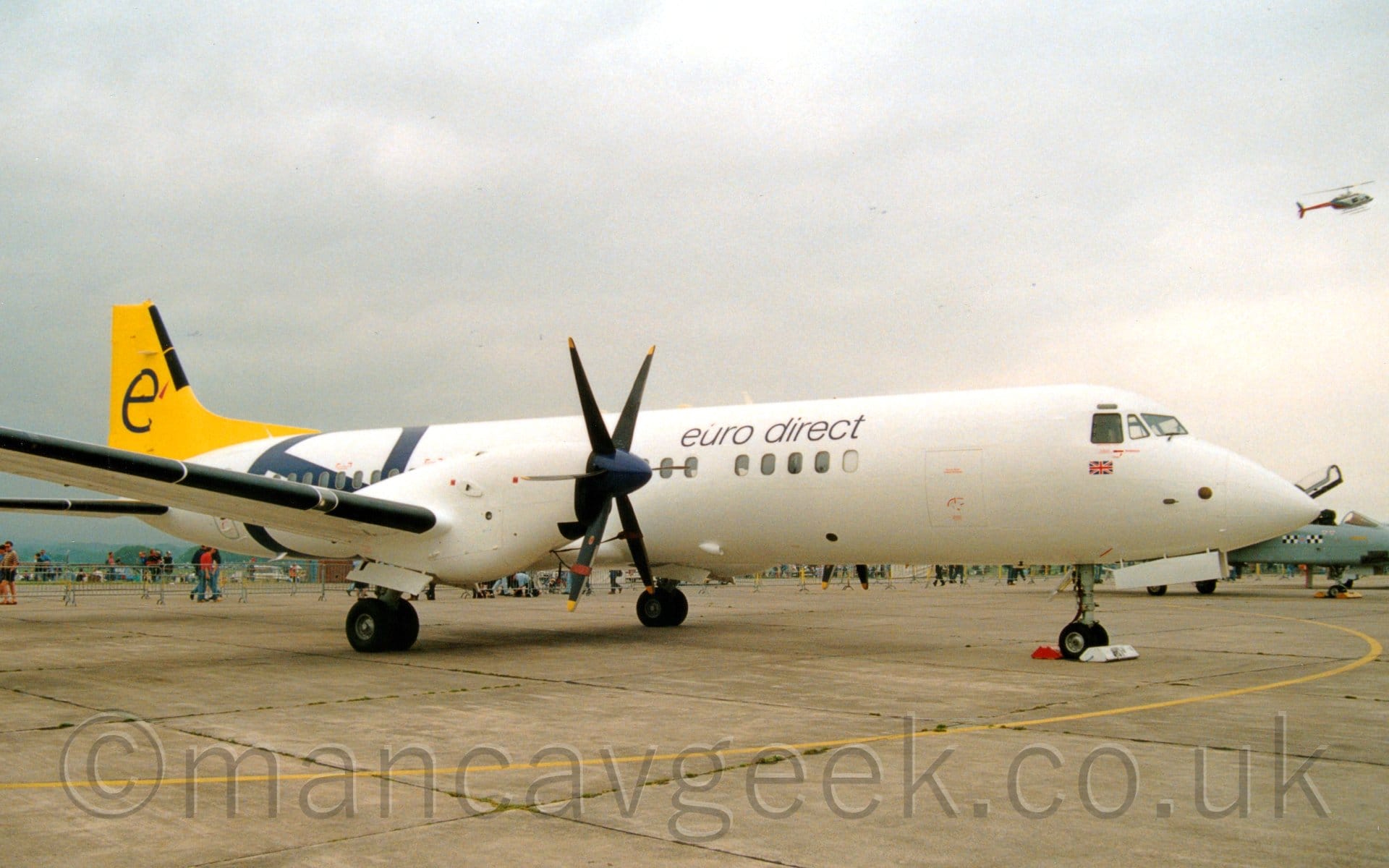 Side view of a twin propellor-engined airliner parked facing to the right. The plane is largely white, with black "Euro Direct" titles on the upper forward fuselage, and a large, lower-case dark blue letter "e" on the rear fuselage. The tail is bright yellow, with a lower-case letter "e" in the middle. Grey concrete apron fills the foreground, with a grey military jet fighter parked in the background on the right, and some people walking past on the left. Grey sky fills the rest of the frame, with a helicopter flying past on the upper right.