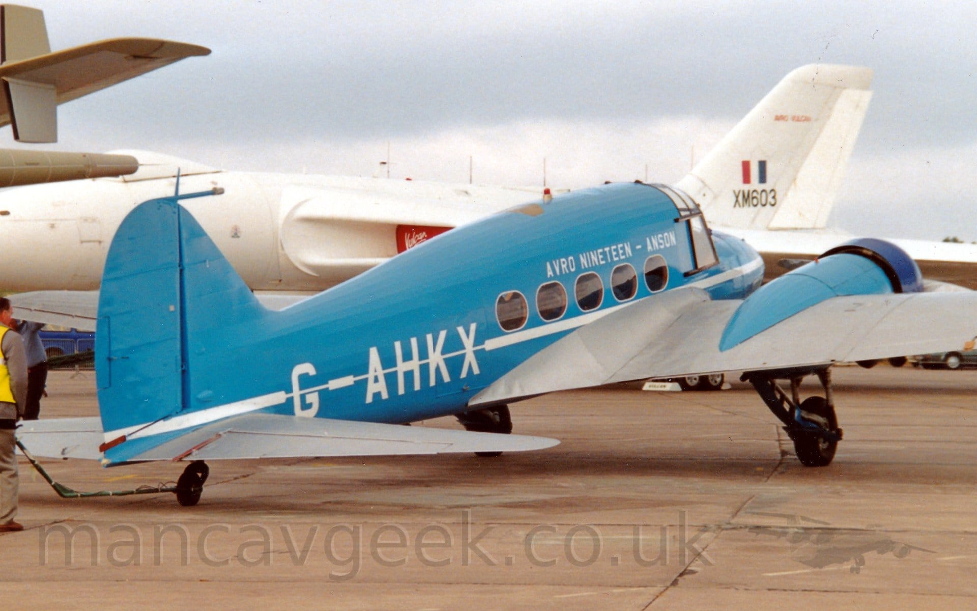 Side view of a twin propellor-engined post-World War 2-era airliner parked on a concrete apron, facing to the right and slightly away from the camera. The plane is mostly blue, with a a thin white stripe running along the body, over-laid with the white registration "G-AHKX" on the rear fuselage, and white text "Avro Nineteen - Anson" on the upper forward fuselage. The wings are an almost metallic grey, while the wing-mounted engine pods, which also house the retractable main landing gear, are the same blue as the fuselage, with the cowling around the engine being a darker blue. The tail of a larger, sandy-brown coloured plane is visible on the left of the frame, while a large, white, 4 jet engined bomber aircraft is parked so if fills the rest of the background, with red engine covers over the engine inlets in the wing roots, and the black serial "XM603"under a red, white, and blue fin flash on the tail. Grey sky completes the rest of the frame.