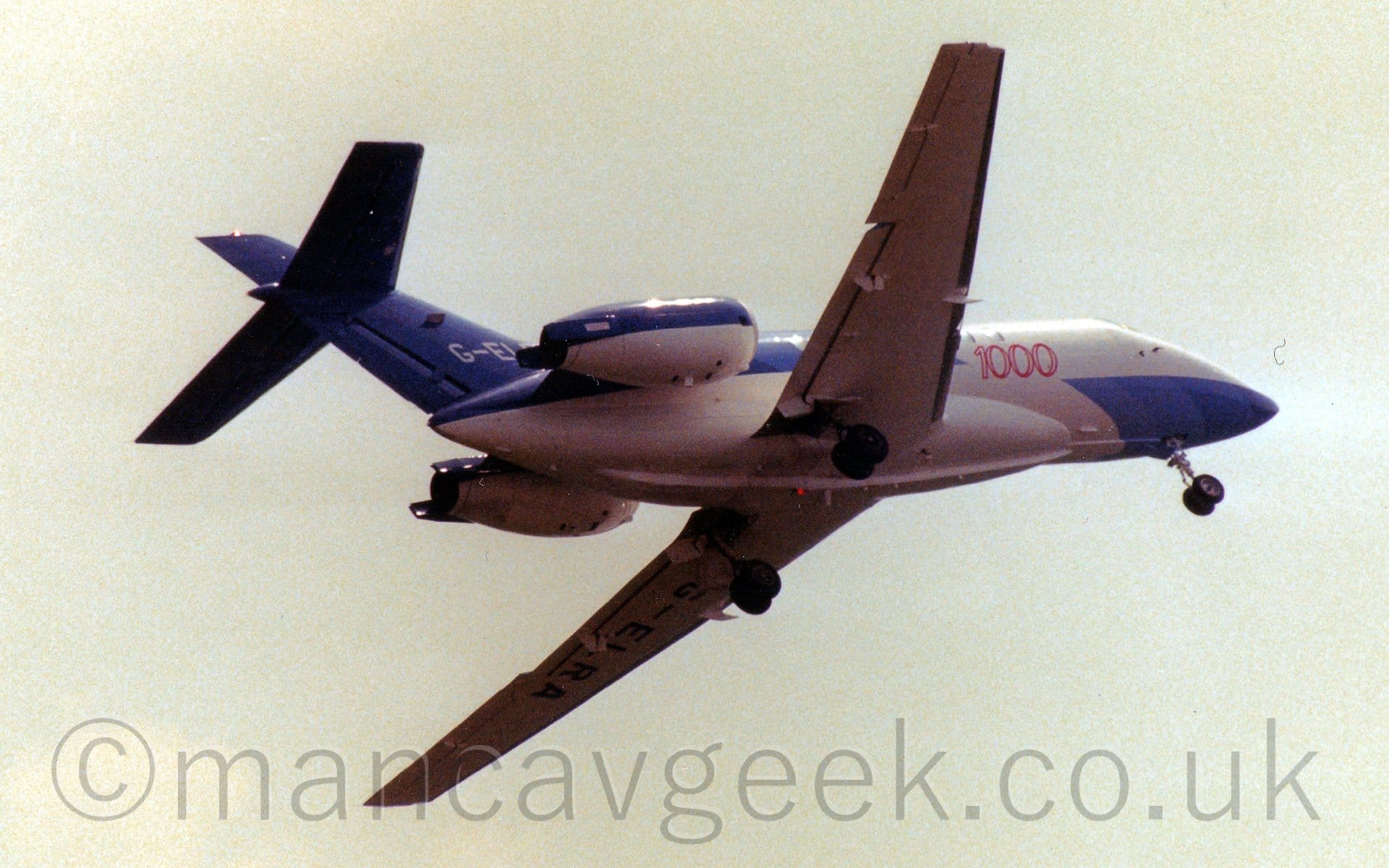 Low side view of a twin engined BizJet flying from left to right at a low altitude. The plane's lower forward fuselage and upper rear fuselage and tail are painted in a geometric pattern made up of various shades of blue, with the rest being white. The red number "1000" is visible on the side of the forward fuselage, while the first 3 letters of the registration, "G-EL" are visible on the tail, the rest obscured by the engines mounted on the sides of the rear fuselage. Bright but hazy grey sky fills the background.