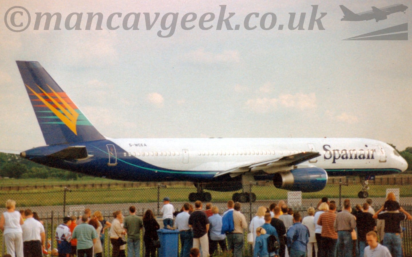 Side view of a twin engined jet airliner taxiing from left to right. The plane is mostly white, with a dark blue belly, rear fuselage, and tail, with a green-blue pinstripe. There are large blue, billboard-style "SpanAir" titles on the forward fuselage, and the black registration "G-MCEA" on the upper rear fuselage. The tail has red, yellow, and greeny-blue spiky shapes, similar to forks, overlaid on each other. The engine pods under the wings are also this dark blue. In the foreground, a large crowd of people are standing in front of a tall, black, chain-link fence, mostly watching this plane. Green grass is visible in the background under the plane, leading up to trees in the distance. Very pale sky with fluffs of white cloud fills the rest of the frame.