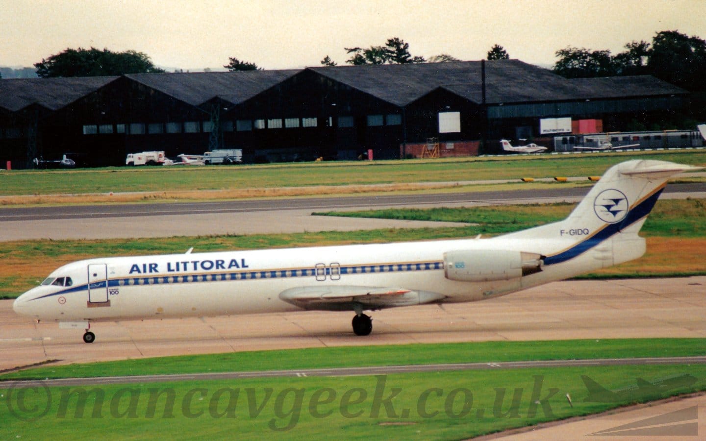 Side view of a twin engined jet airliner with the engines mounted on the sides of the rear fuselage, taxiing from right to left. The plane is almost entirely white, with a blue stripe with a gold pinstripe running along the body, covering the passenger cabin windows, and blue "Air Littoral" titles on the upper forward fuselage. The dark blue registration "F-GIDQ" is on the bottom of the tail, with the blue outline of a circle containing a trio of stylised, blue flying birds. Green grass fills the foreground, with more grass and a runway leading up to black hangars in the distance, with several light aircraft and fuel trucks parked in front. Trees and bright but hazy sky fill the rest of the frame.