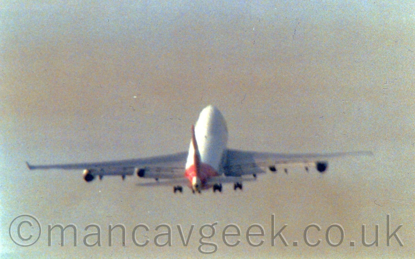 Distant rear view of a very large, 4 engined jet airliner flying away from the camera at a low altitude, with the undercarriage still lowered, flaps extended from the rear of the wings, and the nose raised sharply, suggesting it has just taken off. The plane is mostly white, with a red rear fuselage and tail. The sky was blue, with smears of white cloud, but has been rendered as grey and muddy.