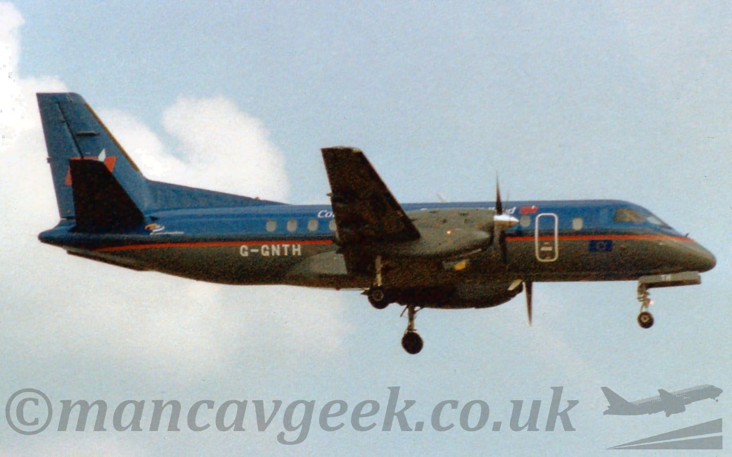 Slightly grainy side view of a twin propellor-engined airliner flying from left to right at a low altitude, with undercarriage lowered and flaps extended from behind the wing, suggesting it is about to land. The plane is mostly dark blue, with a dark grey belly, and a red stripe running along the body. There is white text on the upper fuselage, but only "Co" at the start and "nd" are legible, the rest is being obscured by the wing and engine. There is a red logo with a white diamond on the upper forward fuselage, repeated in a larger form on the tail, and the white registration "G-GNTH" on the lower rear fuselage. Pale blue sky with fluffs of white cloud on the left fill the rest of the frame.