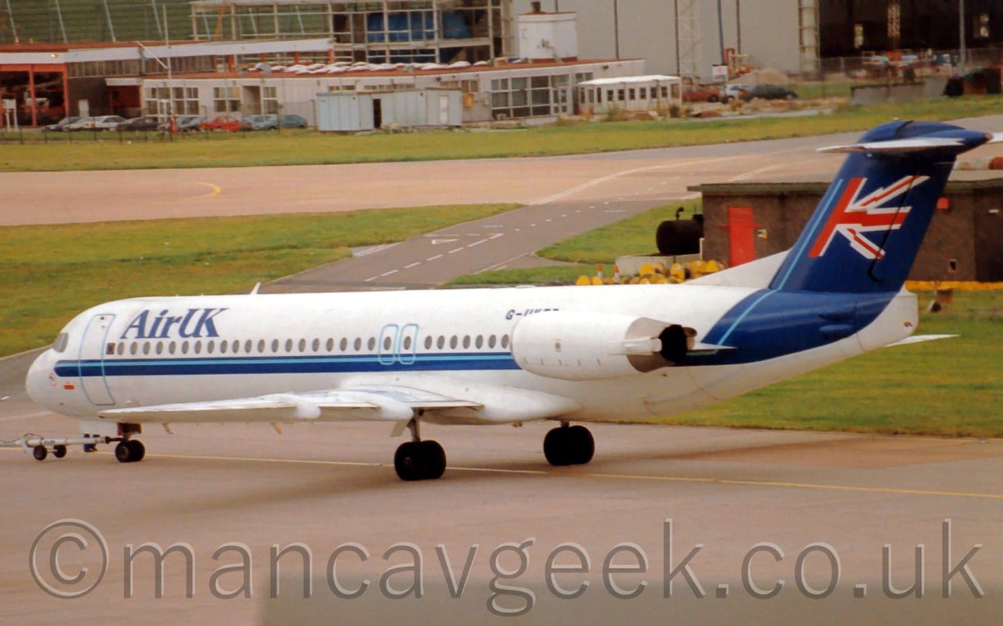 Side view of a twin-engined jet airliner with the engines mounted on the sides of the rear fuselage, facing to the left but being pushed back to the right by an out-of-frame tug attached to the nose-wheel by a long white pole. The plane is mostly white, with a dark and light blue stripe running along the body and sweeping up to fill the tail, where half a British flag is waving on what appears to be a flag pole but is actually that thin light blue stripe. There are dark blue "Air UK" titles on the upper forward fuselage, with the black registration "G-UKFB" on the upper rear fuselage, partially obscured by the white engine pods. Grey concrete apron fills the foreground, with a strip of green grass separating that from a grey taxiway in the distance. There is a brown brick building in the background on the right, directly behind this plane, as well as grey buildings in the far distance.