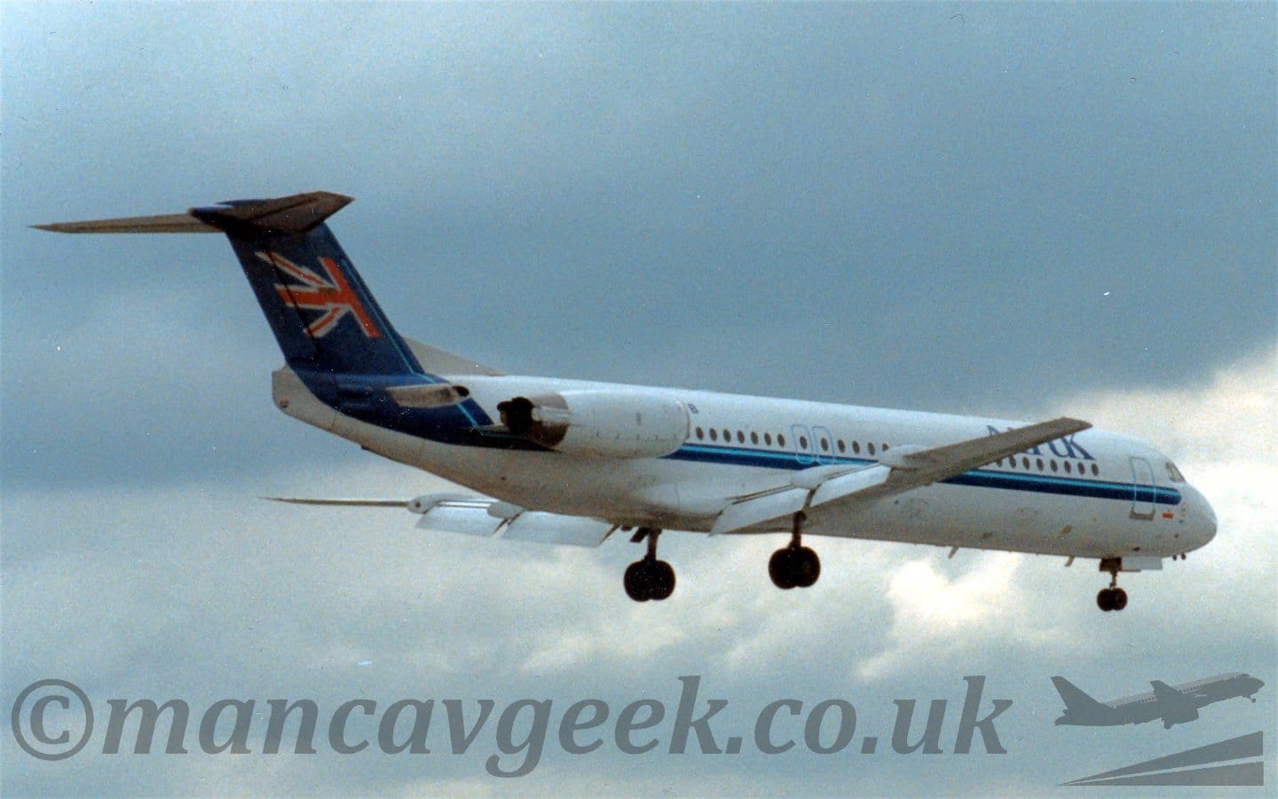 Rear side view of a twin engined jet airliner flying from left to right at a very low altitude, with undercarriage lowered and flaps extended from the rear of the wings, suggesting it is about to land. The plane is mostly white, with a dark and light blue stripe running along the body, before turning up, the dark blue filling the tail, and the light blue part forming a pole for half a fluttering UK flag. There are large, dark blue "Air UK" titles on the upper forward fuselage, while the last letter, "B" of the registration is on the upper rear, the rest of the reg being hidden by the white engines mounted on the sides of the rear fuselage. Dramatic grey skies fill the rest of the frame.