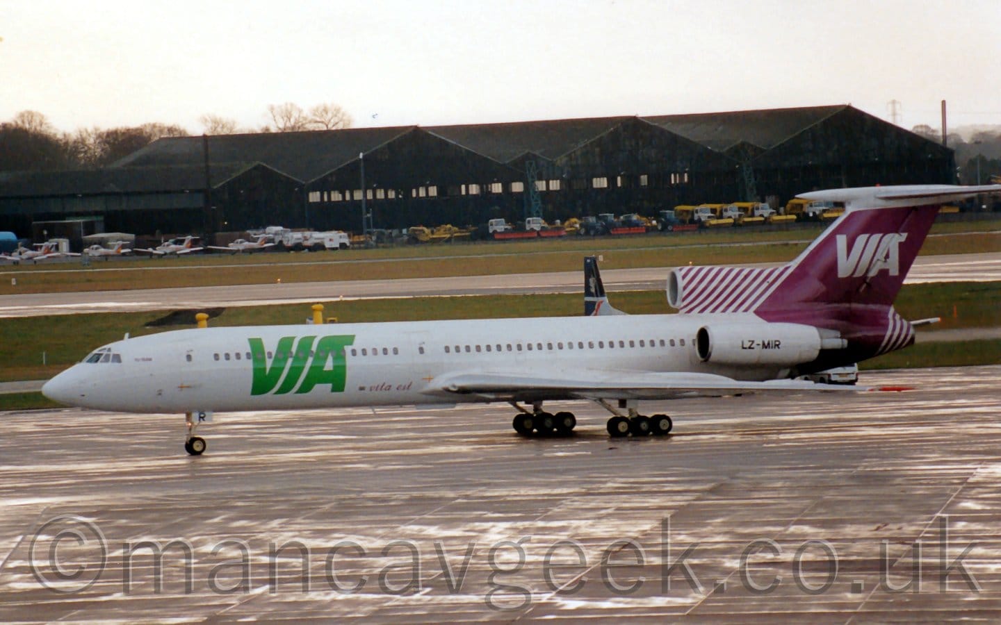 Side view of a 3 engined jet airliner taxiing from right to left. The plane is mostly white, with large, billboard-style green "VIA" titles on the forward fuselage, with much smaller, purple, "Vista Est" at the bottom of the fuselage, just in front of the wings. The tail and rear fuselage are purple, with white "VOA" titles in the middle of the tail, and thin purple and white stripes on the centre engine intake and exhaust in front of and below the tail. The white engines mounted on the sides of the rear fuselage have the black registration "LZ-MIR" on the side. Slightly damp grey concrete apron fills the foreground, with bright sunlight reflecting off smears of water on the ground, while large grassed areas line both sides of a runway in the background. Black hangars with light aircraft and airport vehicles lined up outside stretch across the frame in the distance, under bright but hazy sky.